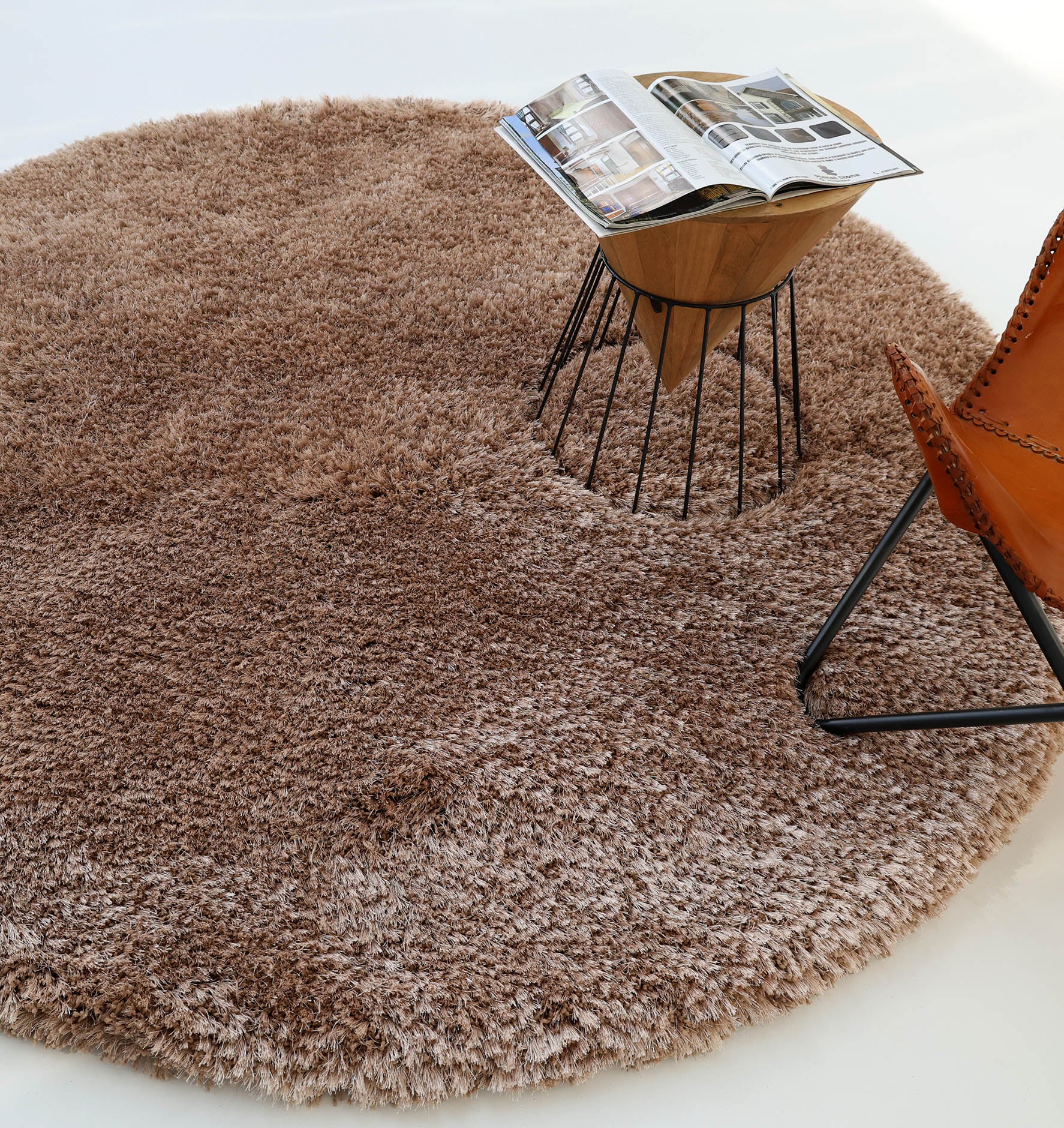 The RAMSHA HOME Soft Plush Brown Shaggy Rug for Living Room - SG 043 sits on a light floor, accompanied by a wooden side table with an open magazine and a leather chair with metal legs.