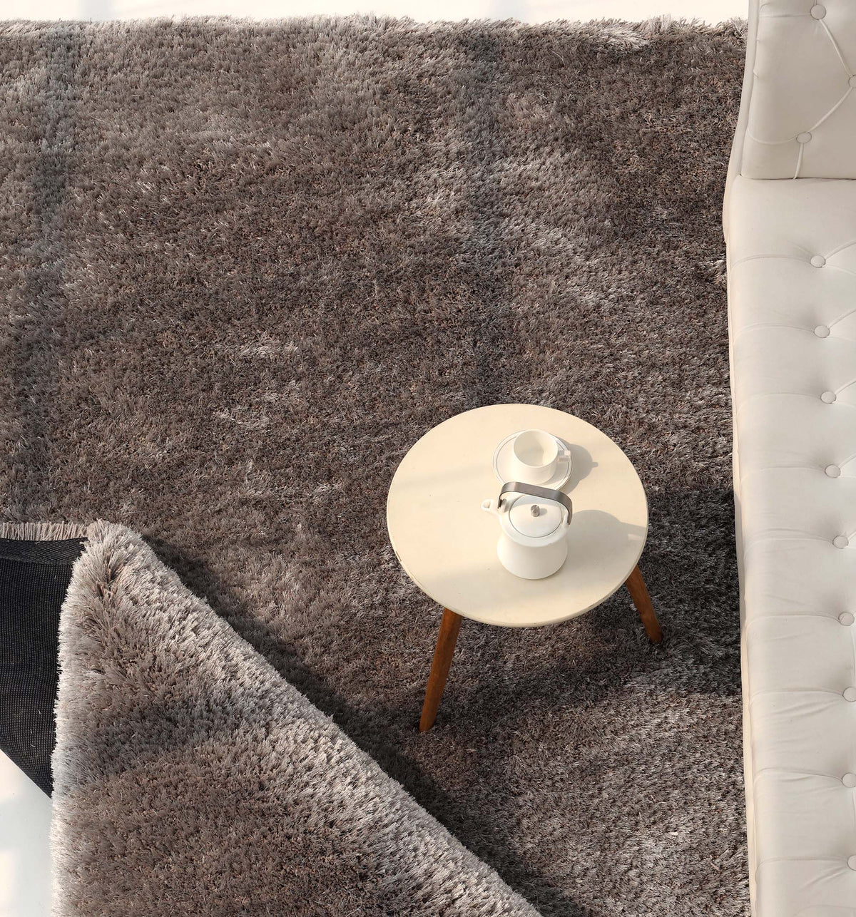 A Soft Plush Grey Shaggy Rug for Living Room - SG 044 by RAMSHA HOME lies slightly folded at one corner next to a white sofa, with a round white table holding a teapot and two cups placed on top.