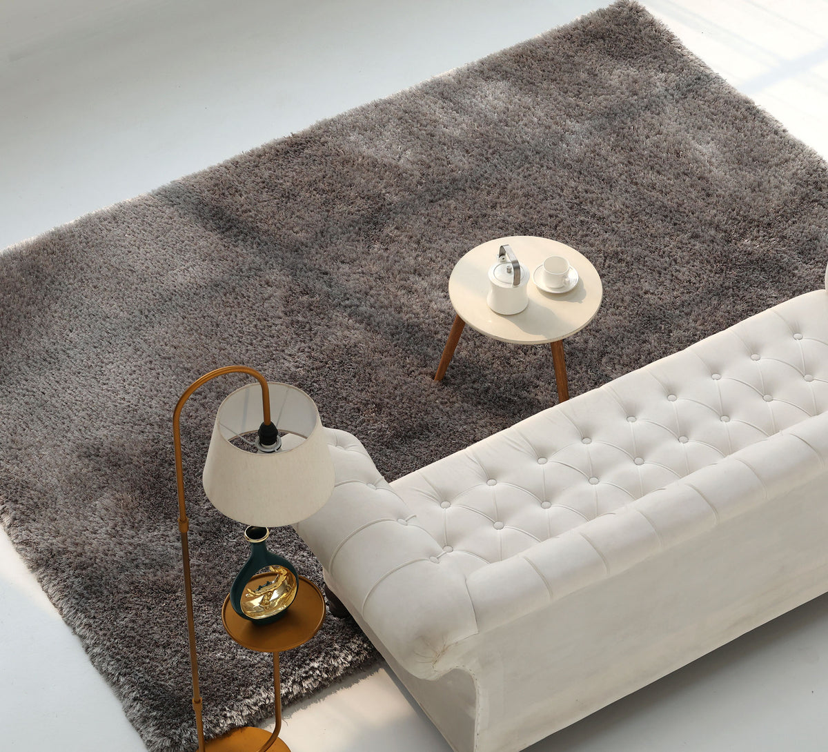 A white tufted sofa sits on a RAMSHA HOME Soft Plush Grey Shaggy Rug for Living Room - SG 044, next to a small round table with a teapot and cups. A floor lamp with a gold base stands nearby, and the scene is well-lit with natural light.