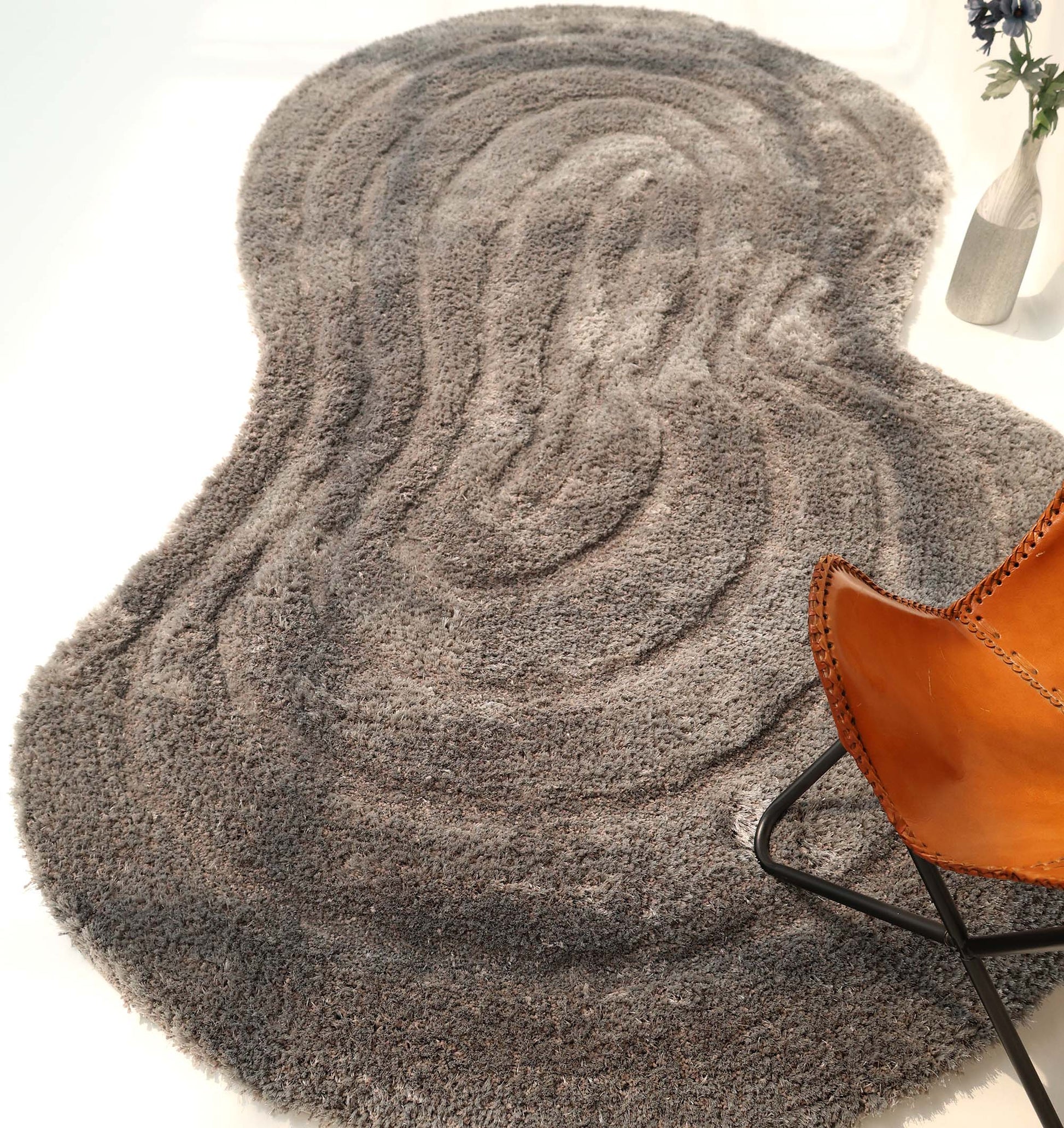 The RAMSHA HOME Luxury Soft Grey Irregular Shaggy Area Rug - SG 044 3D with wavy patterns lies on a white floor, paired with a tan leather chair and a black metal base, while a vase of blue flowers decorates the upper right corner.