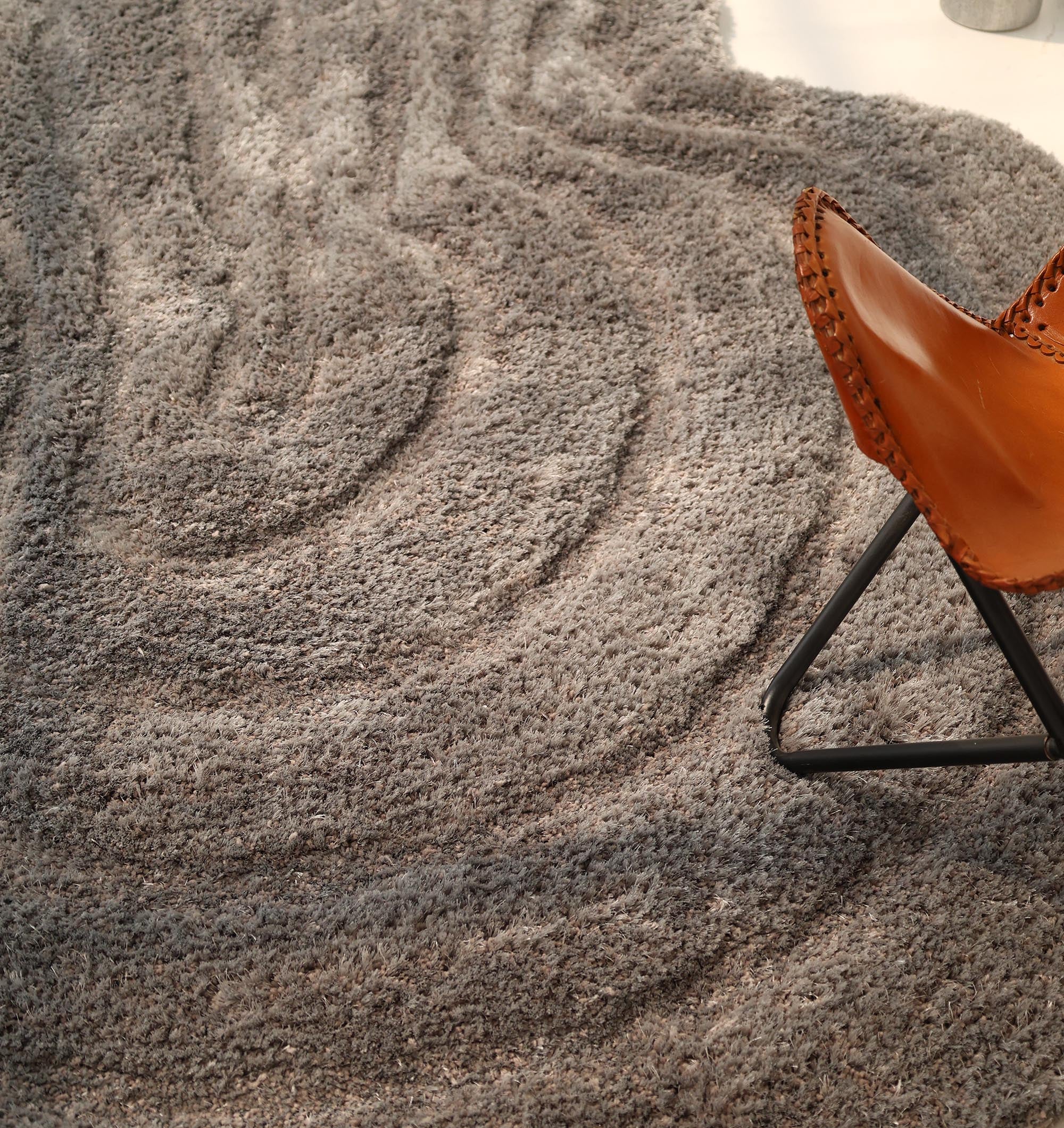 The RAMSHA HOME Luxury Soft Grey Irregular Shaggy Area Rug (SG 044 3D) is displayed beside a brown leather chair with black metal legs. Sunlight highlights the plush grey rug's unique wavy pattern, bringing warmth and style to the space.