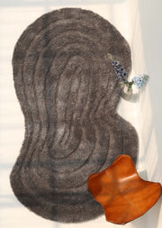 A RAMSHA HOME Luxury Soft Grey Irregular Shaggy Area Rug (SG 044 3D) rests on a light floor with soft shadows; a brown leather chair appears in the lower right, while a small vase with blue and white flowers sits near the top right edge.