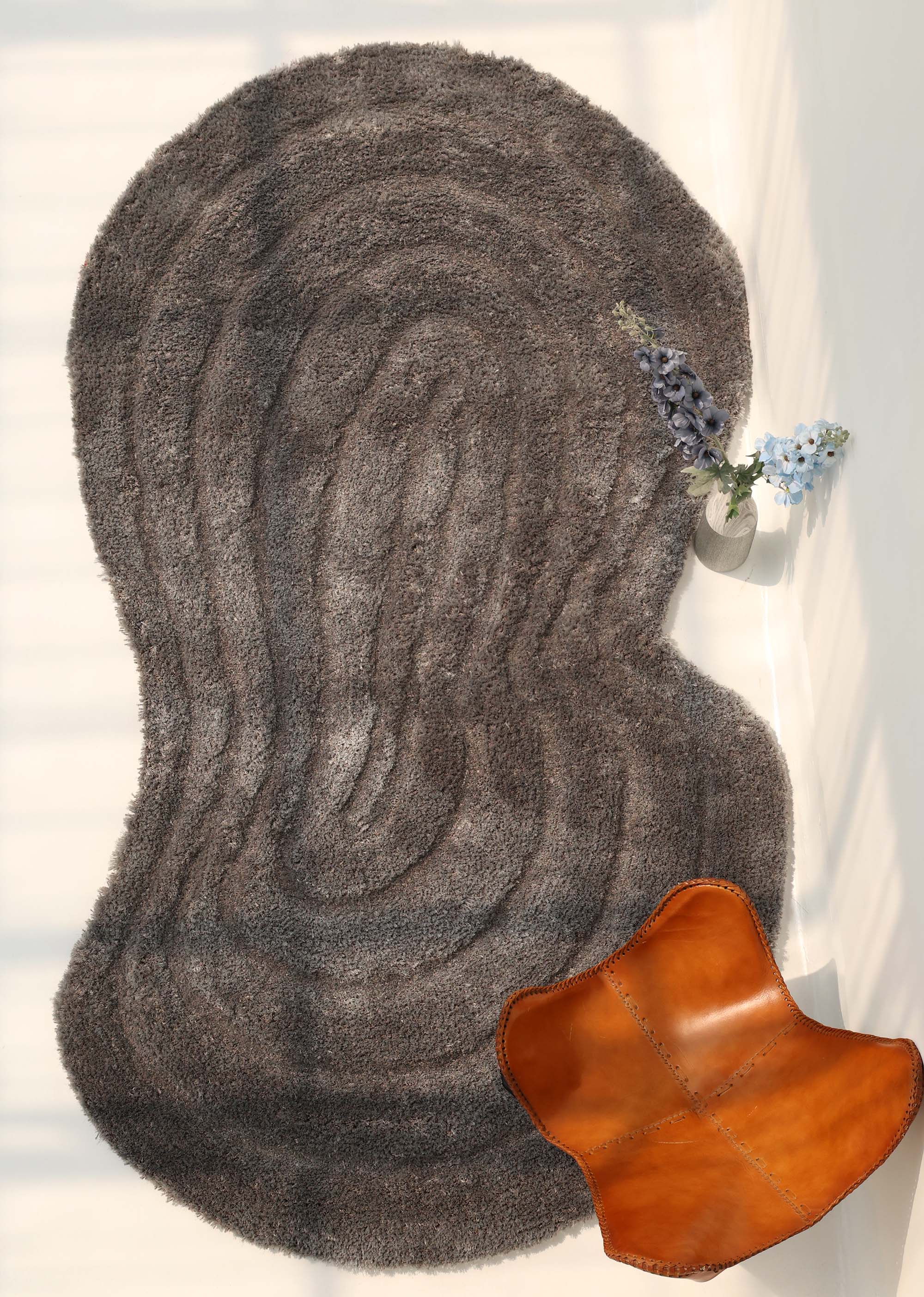 A RAMSHA HOME Luxury Soft Grey Irregular Shaggy Area Rug (SG 044 3D) rests on a light floor with soft shadows; a brown leather chair appears in the lower right, while a small vase with blue and white flowers sits near the top right edge.