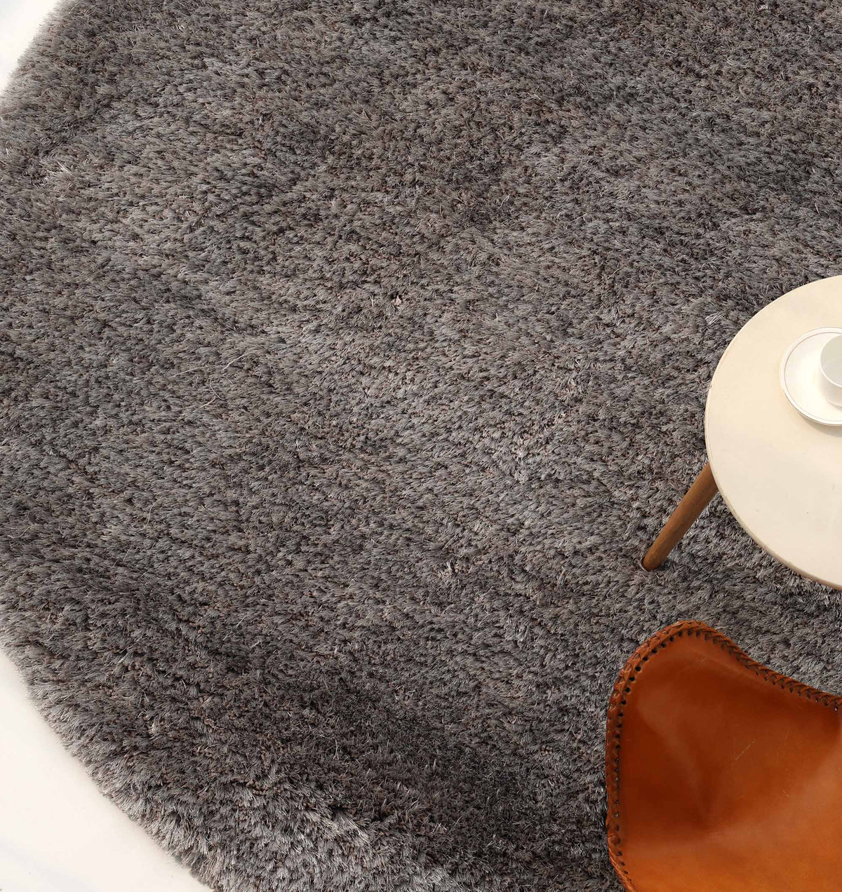 A soft, plush grey shaggy rug from RAMSHA HOME (SG 044) lies on the floor with a small white table placed on top, holding a cup and saucer. A brown leather chair is partially visible near the table.