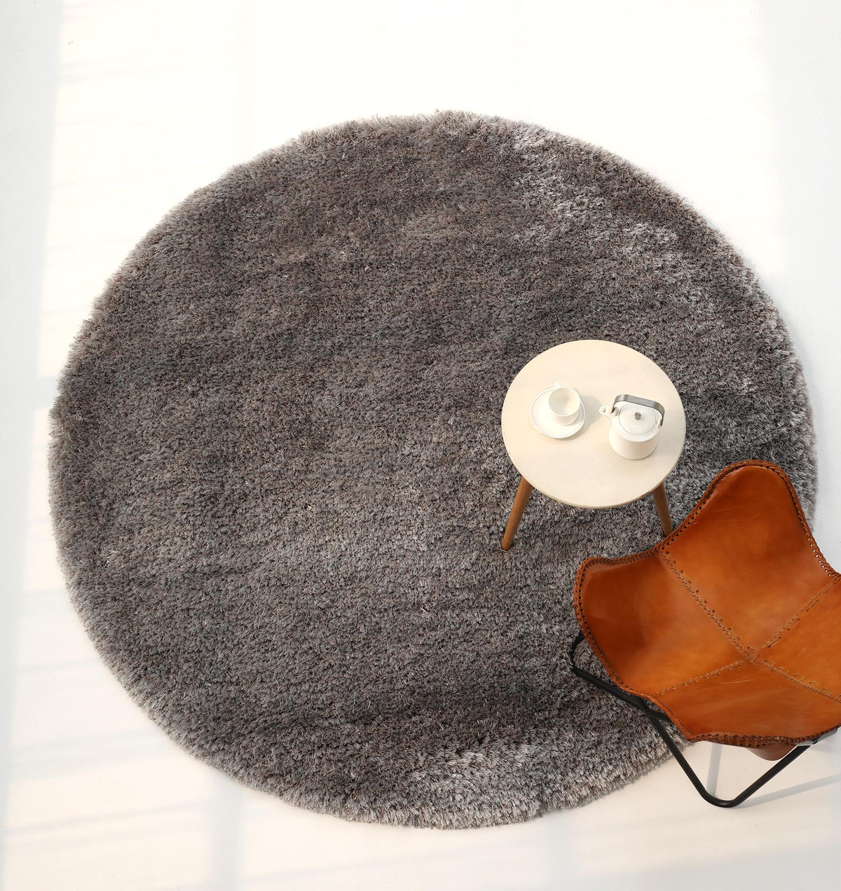 A RAMSHA HOME Soft Plush Grey Shaggy Rug for Living Room (SG 044) sits beneath a small round table holding two white cups, next to a brown leather chair, viewed from above in a brightly lit room.