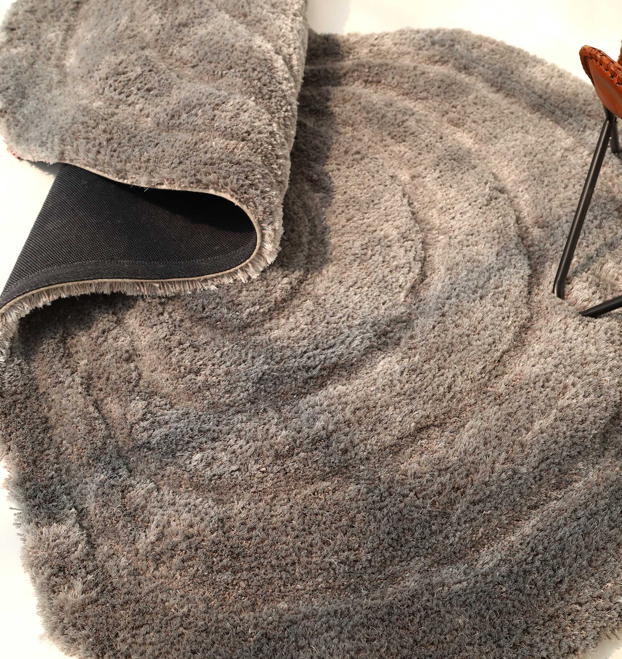 The RAMSHA HOME Luxury Soft Grey Irregular Shaggy Area Rug - SG 044 3D features a gray, circular, wavy-textured design. The image shows part of the rug flipped to reveal its black underside and a chair's corner on the right.