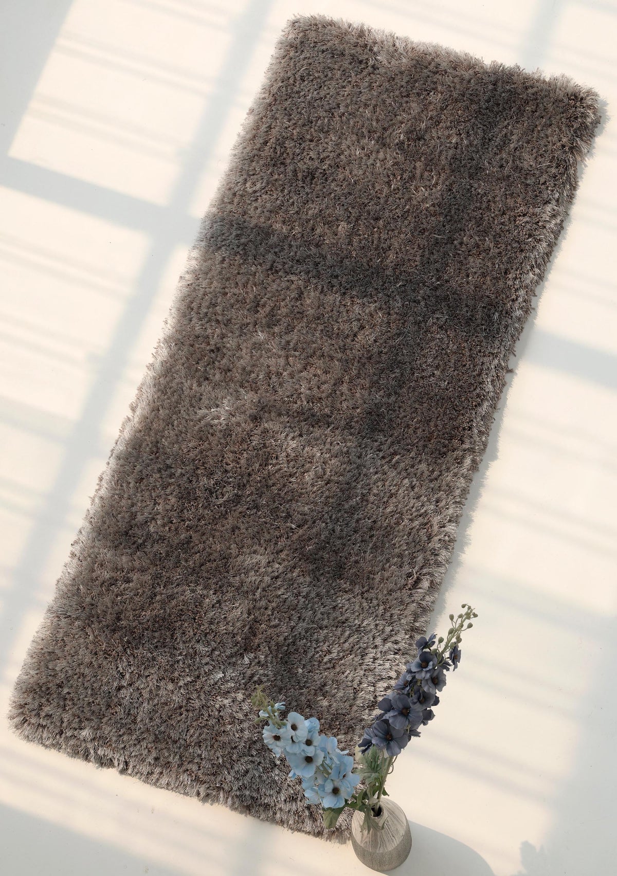 A rectangular grey Soft Plush Shaggy Rug for Living Room by RAMSHA HOME (SG 044) is placed on a light floor with sunlight streaming in, casting gentle shadows. A small vase with blue and purple flowers sits near one corner of the rug.