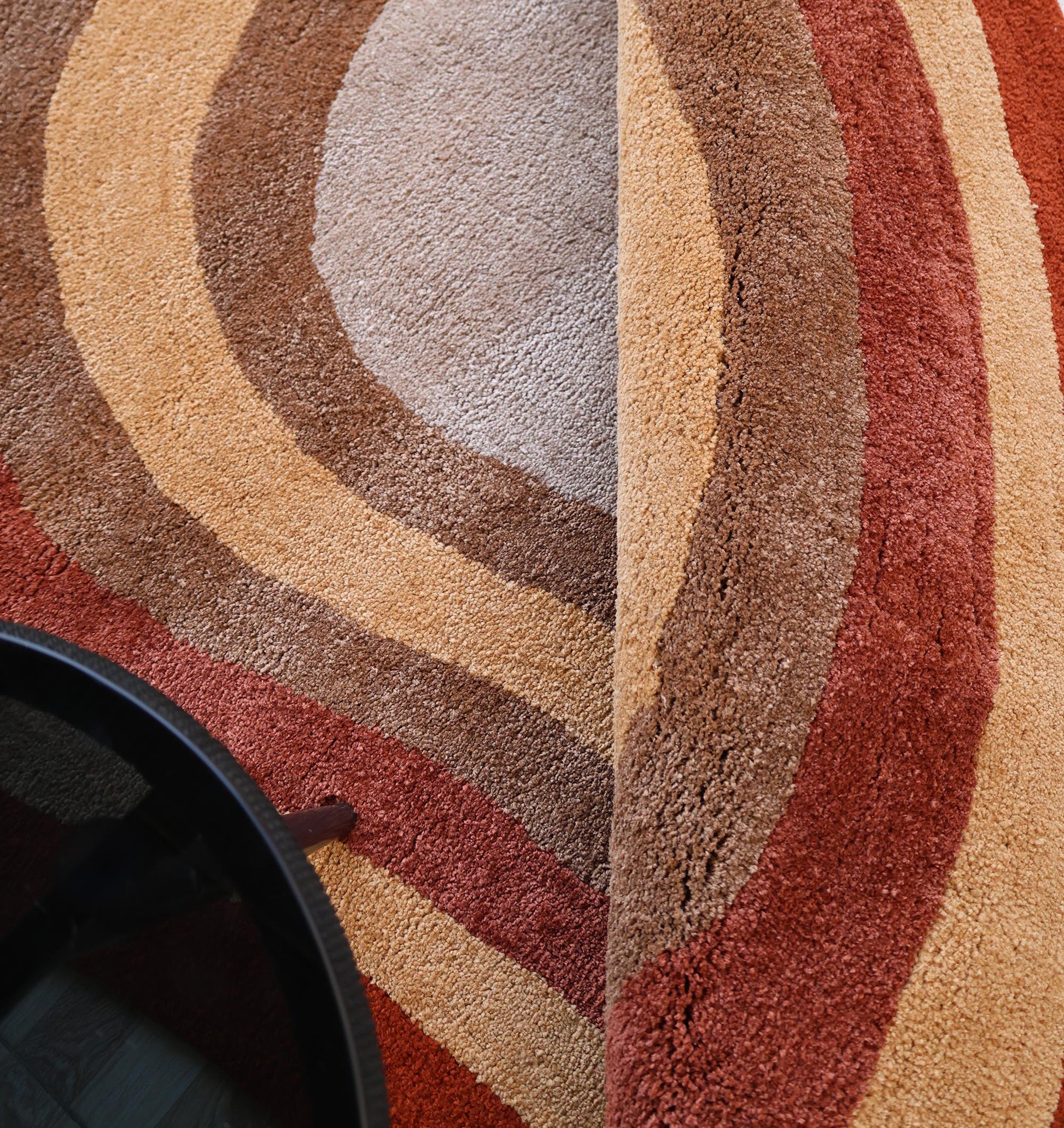 A close-up of the RAMSHA HOME Soft Plush Abstract Shape Shaggy Rug – Terracotta Beige- SG 045 reveals wavy cream, yellow, brown, and burnt orange stripes. A black round table in the corner highlights this modern rug’s retro appeal.