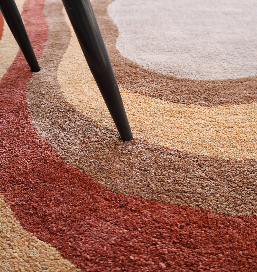 Close-up of RAMSHA HOME’s Soft Plush Abstract Shape Shaggy Rug – Terracotta Beige- SG 045, featuring an abstract layered design in red, brown, beige, and tan. A black metal furniture leg stands on the plush surface, casting a slight shadow.