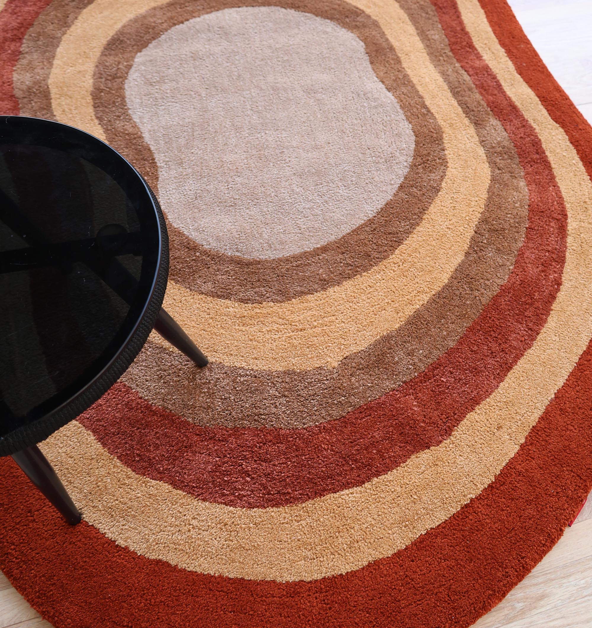 A black round side table sits on the RAMSHA HOME Soft Plush Abstract Shape Shaggy Rug – Terracotta Beige- SG 045, featuring an abstract layered design in beige, brown, tan, and rust tones.