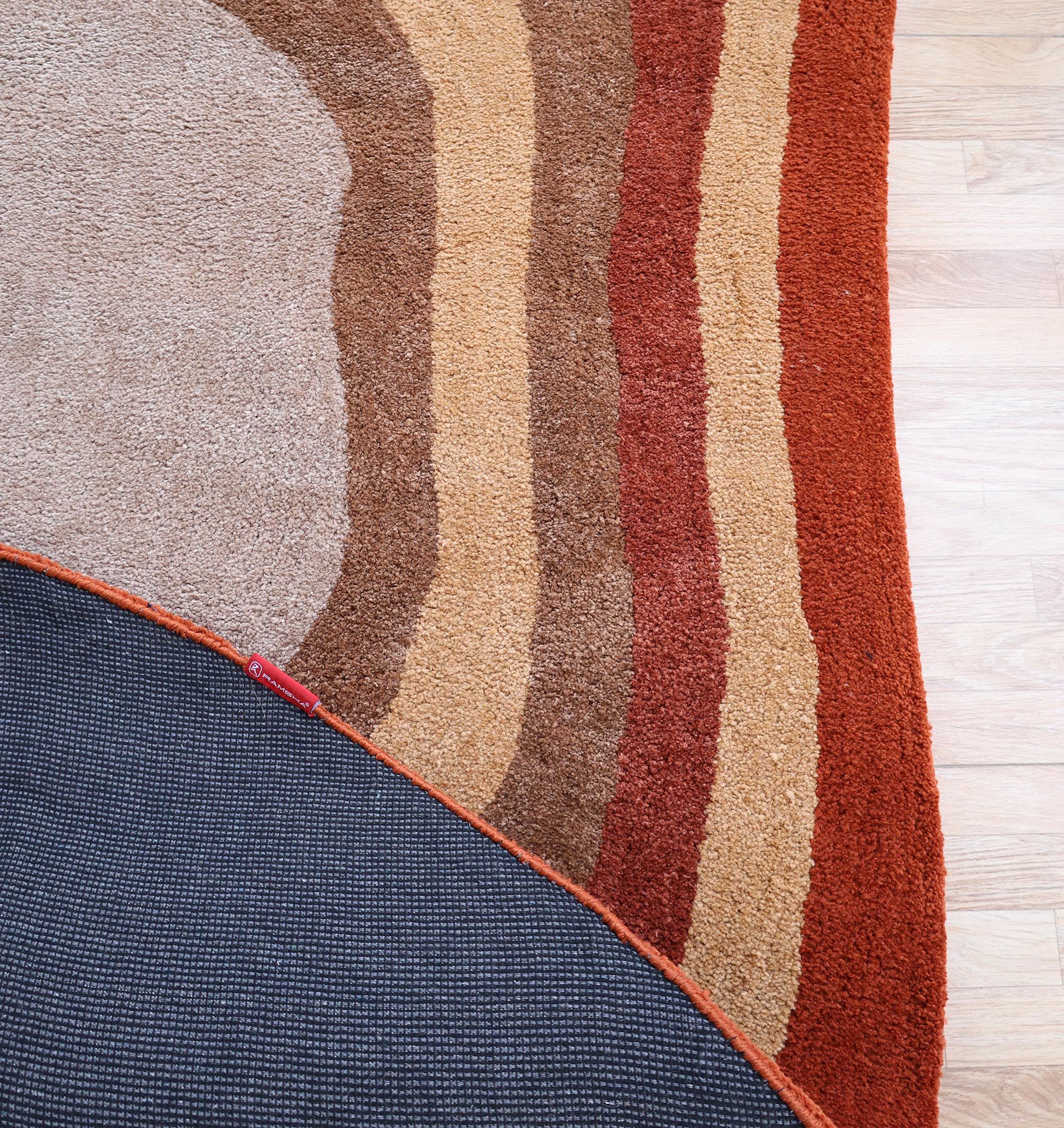 The RAMSHA HOME Soft Plush Abstract Shape Shaggy Rug – Terracotta Beige (SG 045) features curved brown, tan, and red stripes on a wooden floor, with a flipped corner showing its dark, textured non-slip backing.