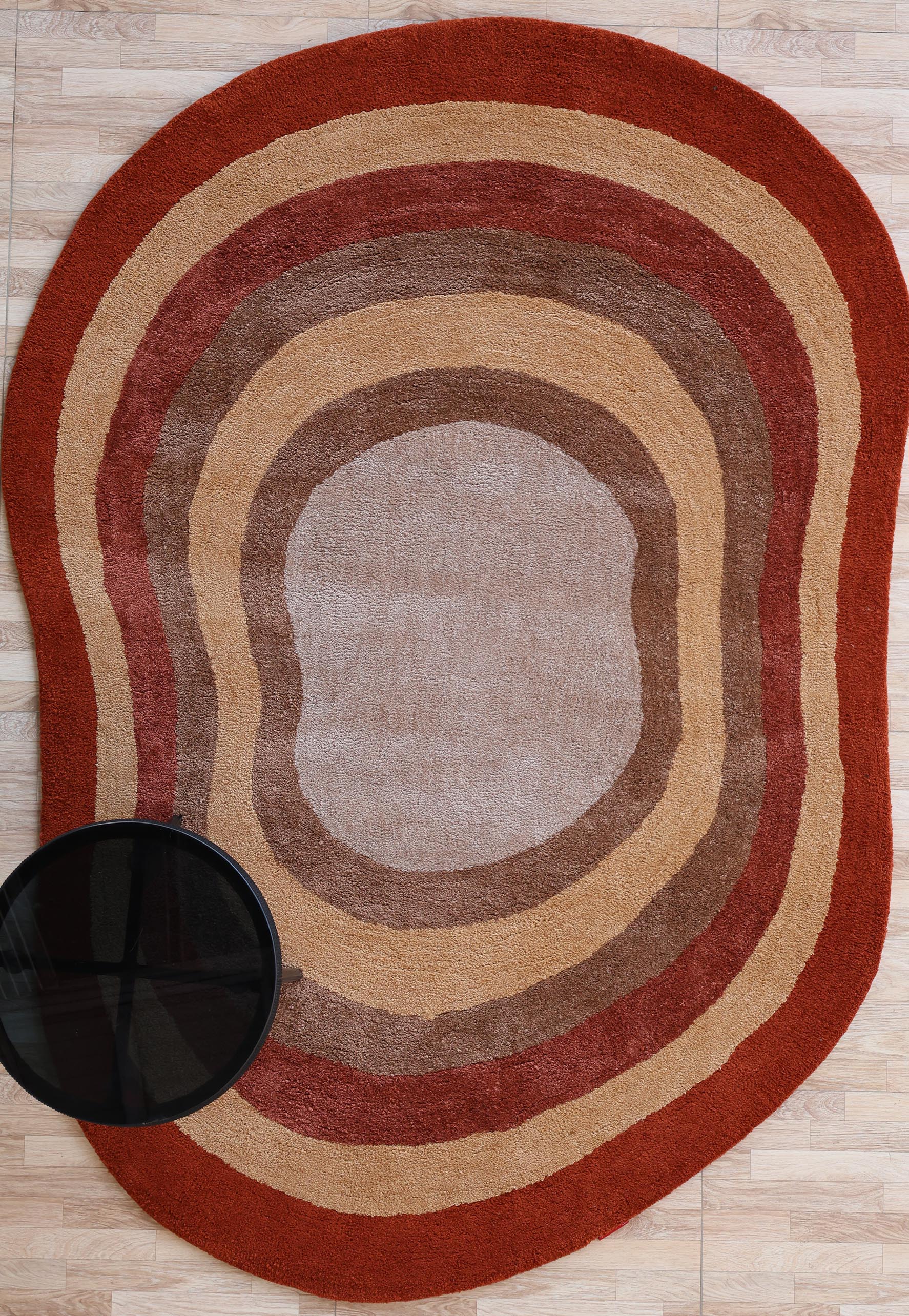 The RAMSHA HOME Soft Plush Abstract Shape Shaggy Rug – Terracotta Beige- SG 045, with layered oval designs in red, brown, tan, and beige, lies on a light wood floor. A round black side table sits partially atop the rug in the bottom left corner.