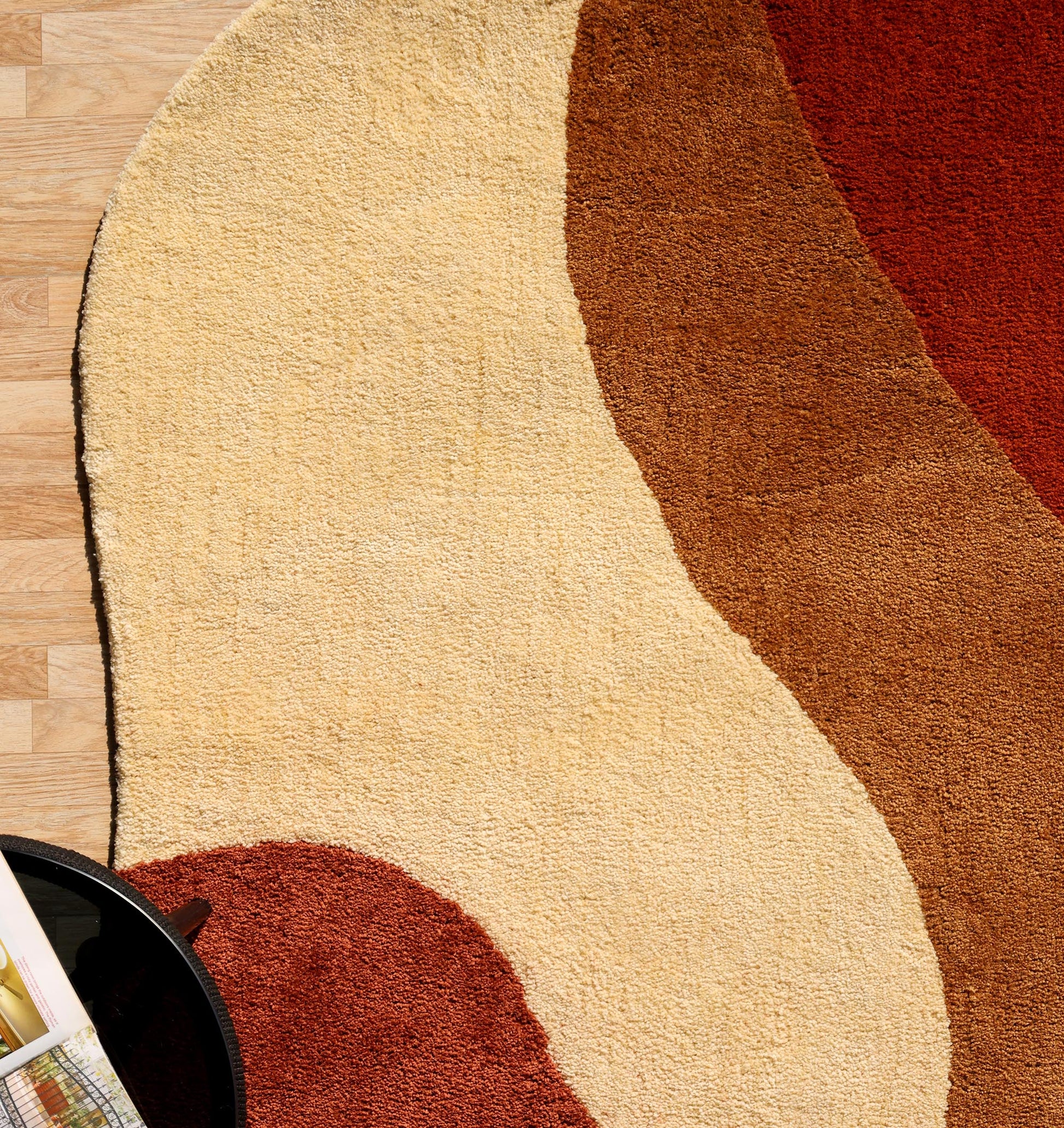 The RAMSHA HOME Terracotta Beige Soft Irregular Shape Shaggy Rug (SG 046) with wavy beige, brown, and rust tones is placed on a light wood floor. The lower left shows part of a round black table holding a magazine.