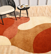 A cozy, modern room features the RAMSHA HOME Terracotta Beige Soft Irregular Shape Shaggy Rug (SG 046) layered on a wooden floor, with a round black chair, beige cushion, and a small black table holding an open book nearby.