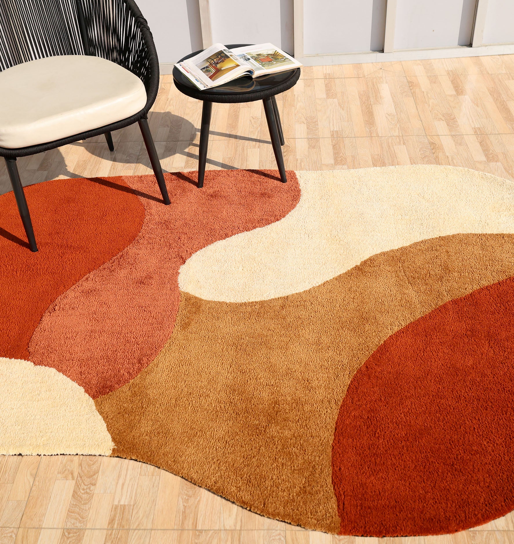 A cozy, modern room features the RAMSHA HOME Terracotta Beige Soft Irregular Shape Shaggy Rug (SG 046) layered on a wooden floor, with a round black chair, beige cushion, and a small black table holding an open book nearby.