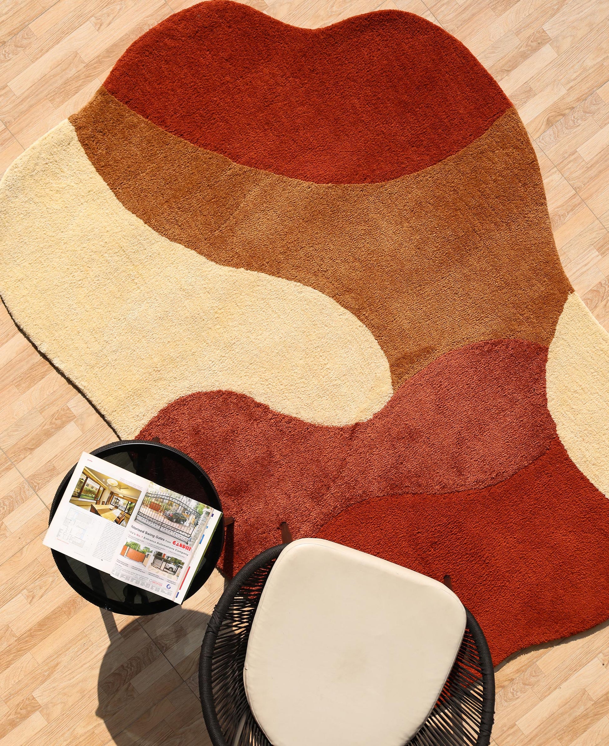 The RAMSHA HOME Terracotta Beige Soft Irregular Shape Shaggy Rug (SG 046) lies on a wooden floor beside a black side table with an open magazine and a chair with a light-colored cushion.
