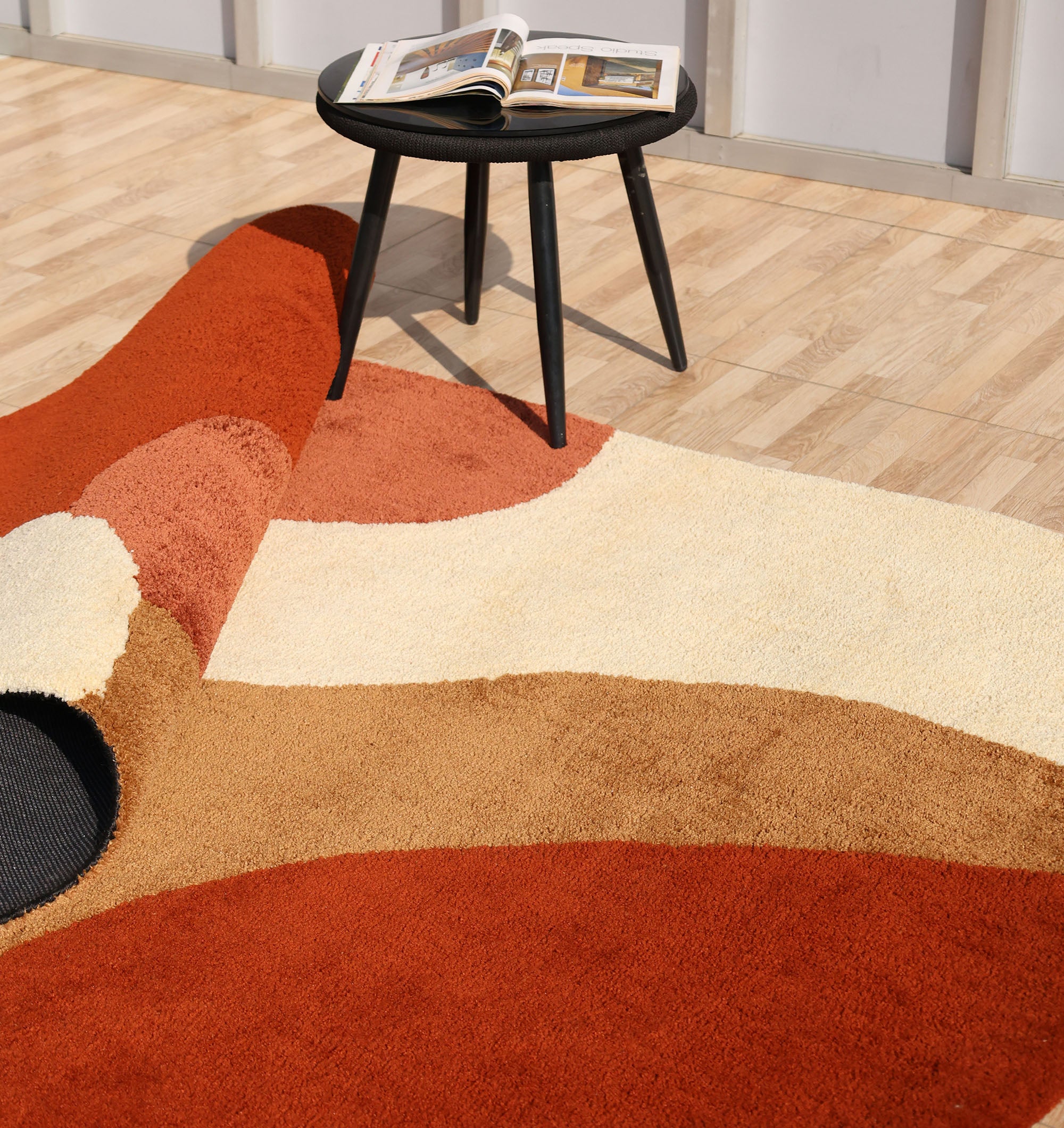 The RAMSHA HOME Terracotta Beige Soft Irregular Shape Shaggy Rug (SG 046) in cream, brown, and orange is partly folded on a light wood floor beside a black round table holding an open magazine.