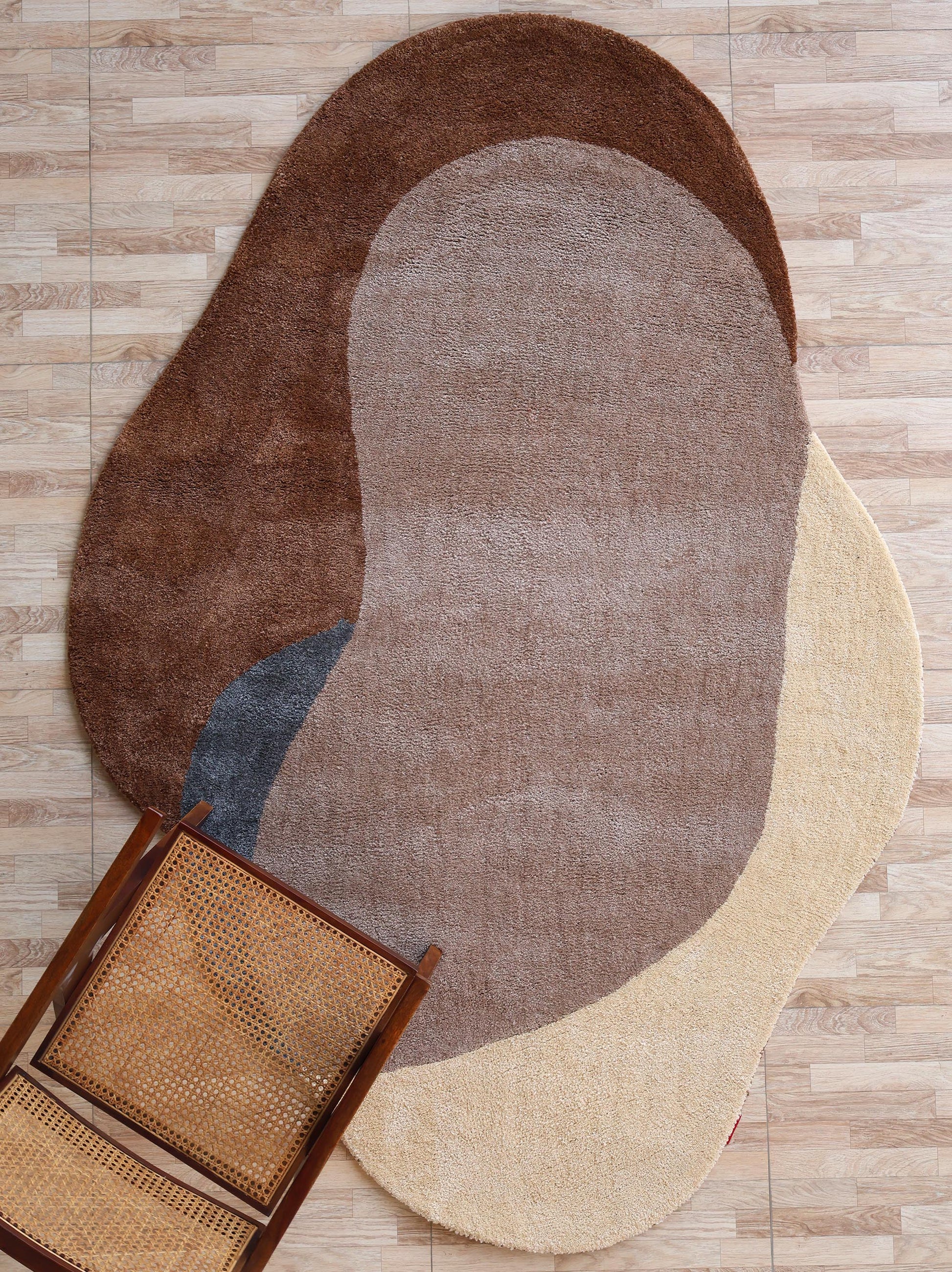 The RAMSHA HOME Contemporary Freeform Plush Rug - SG 047, featuring overlapping brown, beige, tan, and gray shapes, lies on a light wood floor with a wooden chair and woven cane seat in the lower left corner.