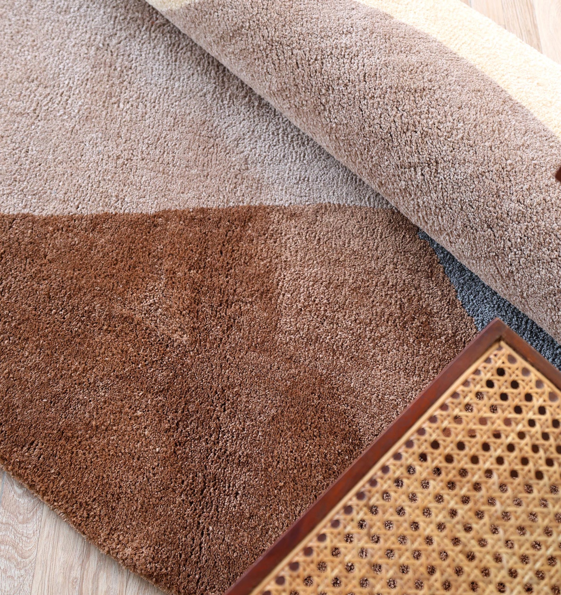 A close-up of the RAMSHA HOME Contemporary Freeform Plush Rug - SG 047, rolled up in brown and beige tones on a light wooden floor, with part of a cane-seated wooden chair in the foreground.