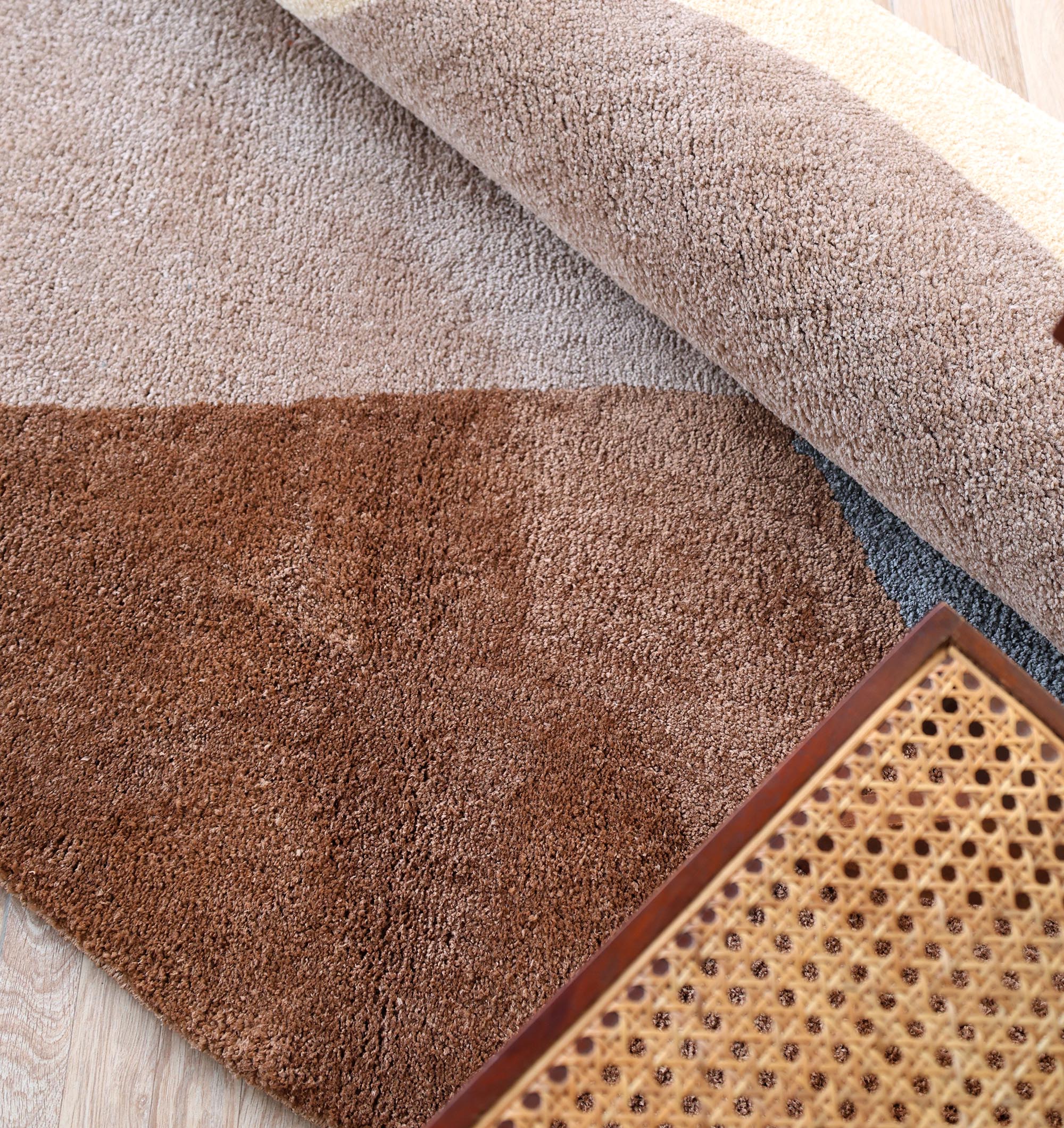 A close-up of the RAMSHA HOME Contemporary Freeform Plush Rug - SG 047, rolled up in brown and beige tones on a light wooden floor, with part of a cane-seated wooden chair in the foreground.