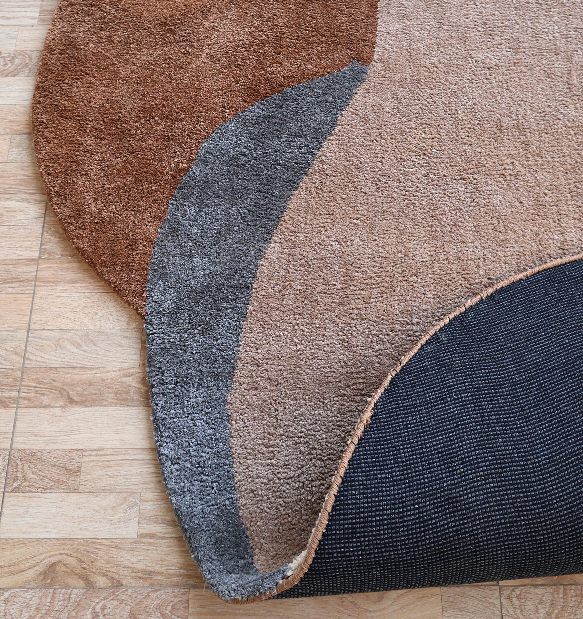 A close-up of the RAMSHA HOME Contemporary Freeform Plush Rug - SG 047, featuring soft beige, brown, and gray sections on a wood floor with one corner lifted to reveal its black textured backing.