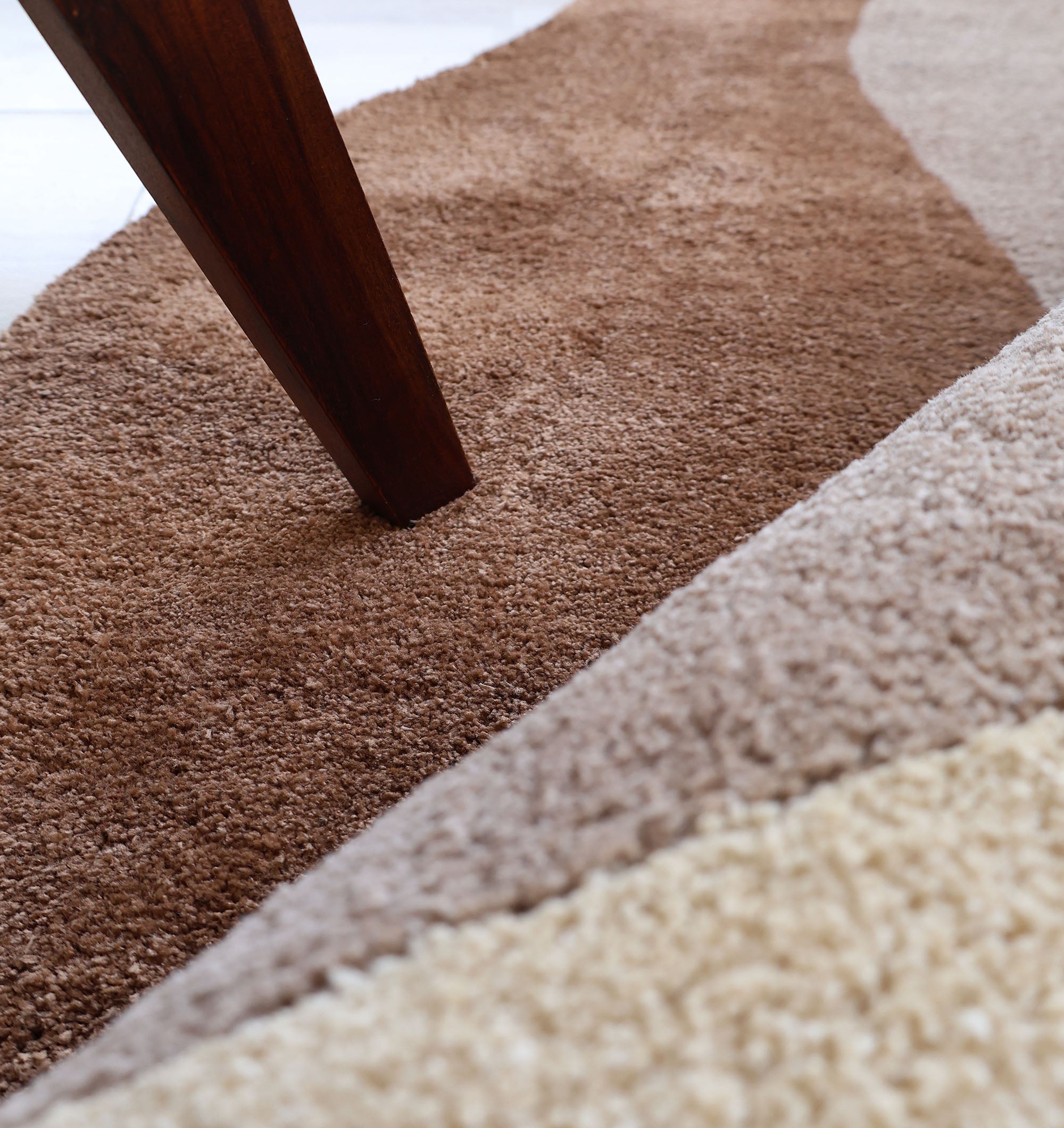 A close-up of a wooden furniture leg on the RAMSHA HOME Contemporary Freeform Plush Rug - SG 047, featuring a wavy, textured multicolored design in brown, beige, and tan shades.