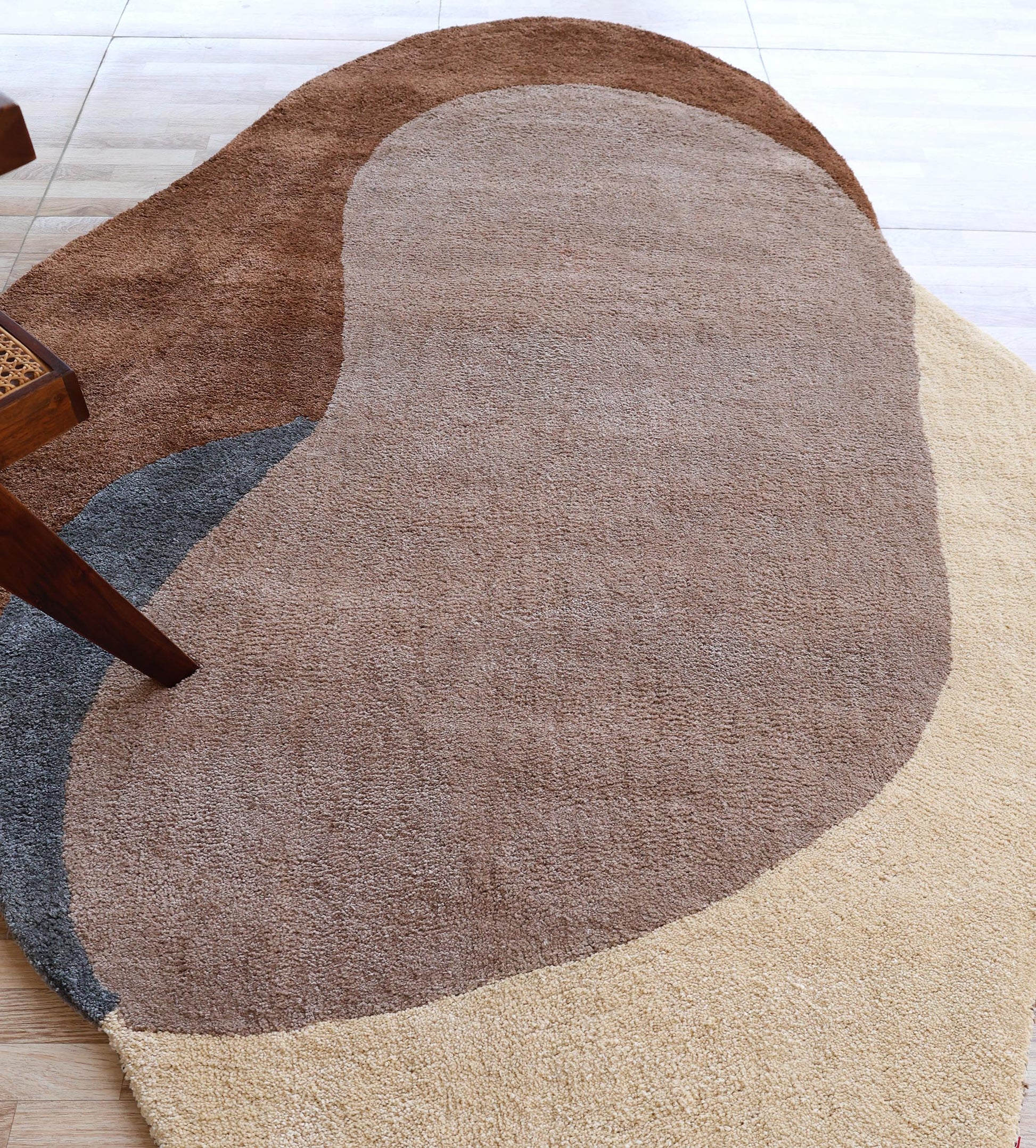 The RAMSHA HOME Contemporary Freeform Plush Rug - SG 047, featuring overlapping beige, brown, tan, and dark gray sections in an abstract kidney shape, lies on a light wood floor with its plush shaggy texture beside a wooden chair leg.