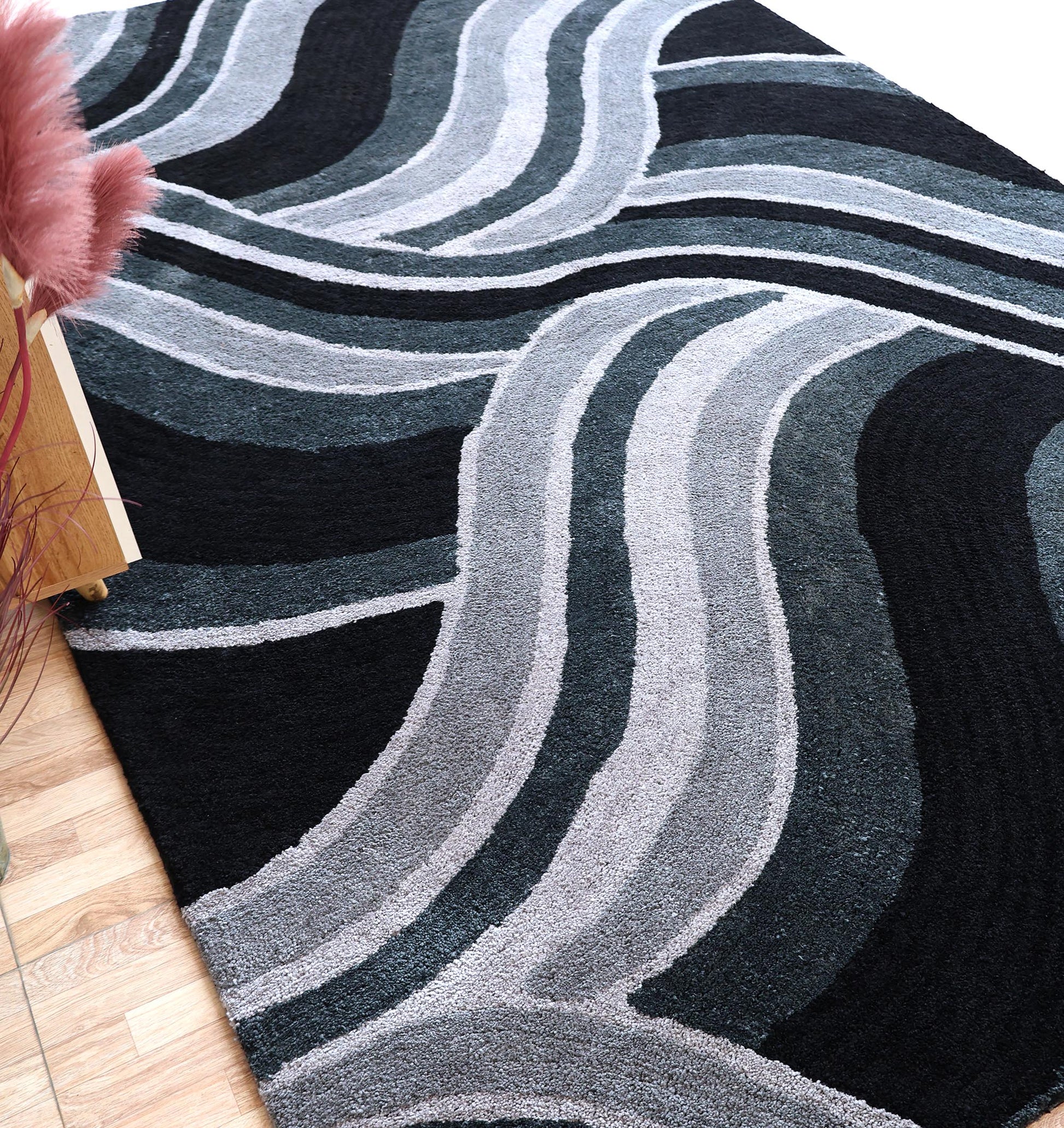 The RAMSHA HOME Multi Shaggy Area Rug for Large Living Room - SG 048 features wavy, overlapping lines in black, gray, and white, shown on a hardwood floor beside a pink plant and wooden table.