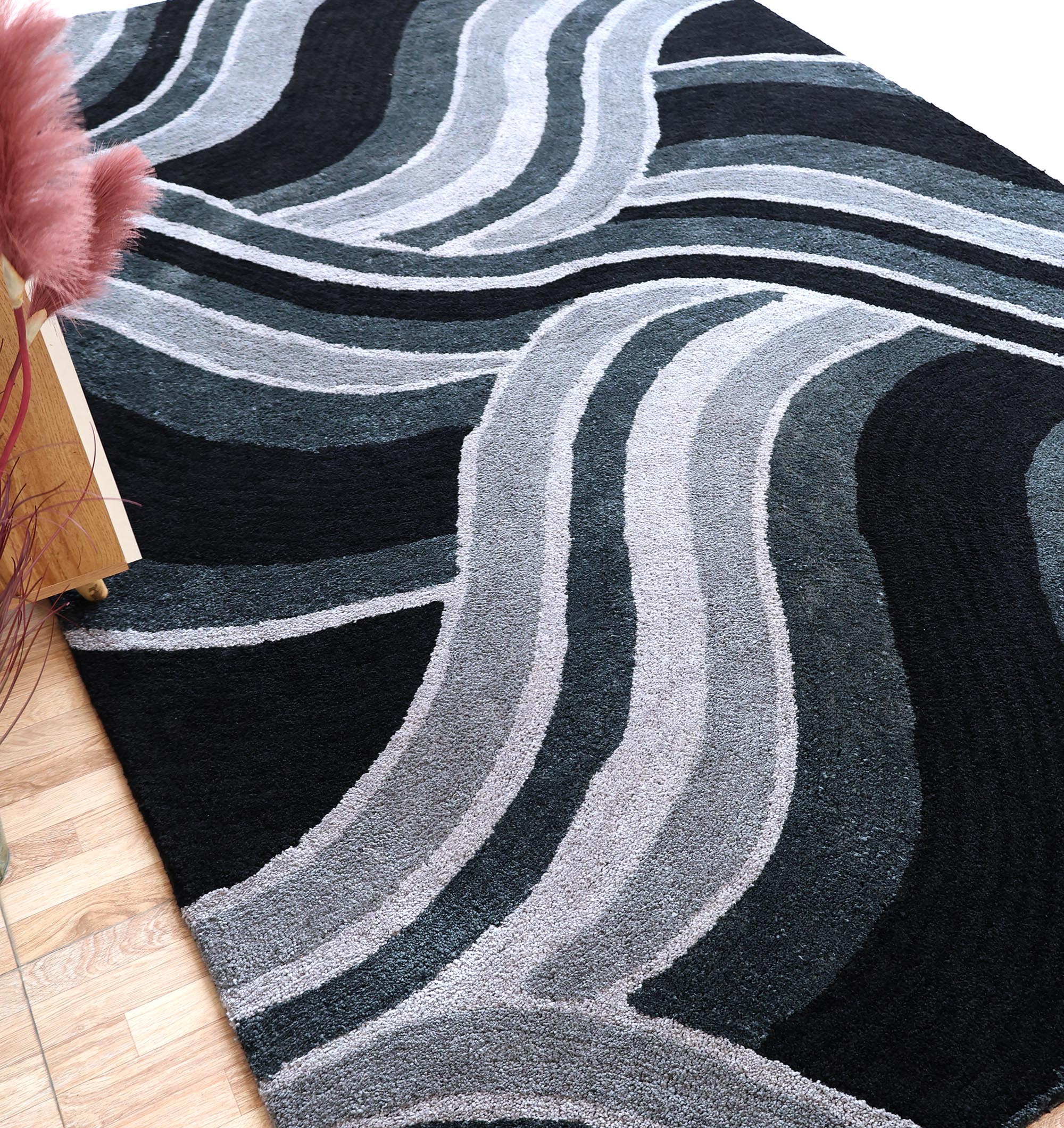 The RAMSHA HOME Multi Shaggy Area Rug for Large Living Room - SG 048 features wavy, overlapping lines in black, gray, and white, shown on a hardwood floor beside a pink plant and wooden table.
