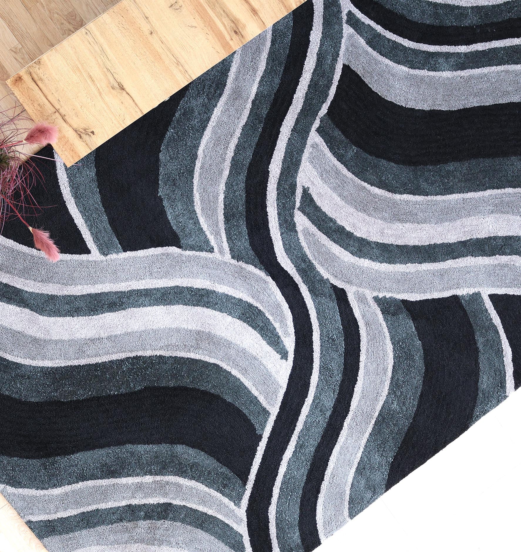 The RAMSHA HOME Multi Shaggy Area Rug for Large Living Room (SG 048) features modern wavy abstract lines in black, gray, and white, shown partially on a light wood floor with pink decorative plant stems in the corner.