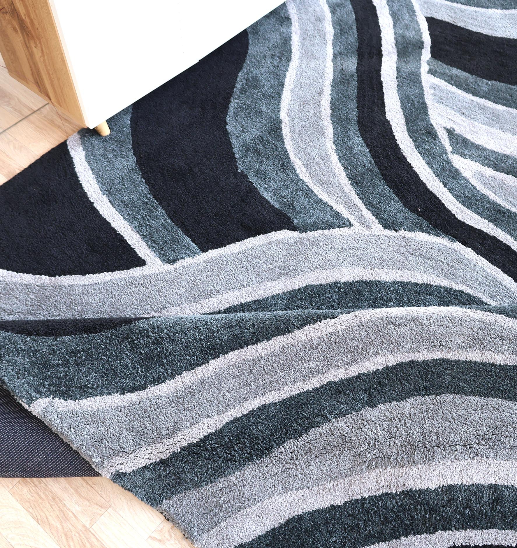 A close-up of the RAMSHA HOME Multi Shaggy Area Rug for Large Living Room - SG 048, featuring wavy patterns in gray, black, and blue, partially lifted at one corner on a wooden floor beside furniture.