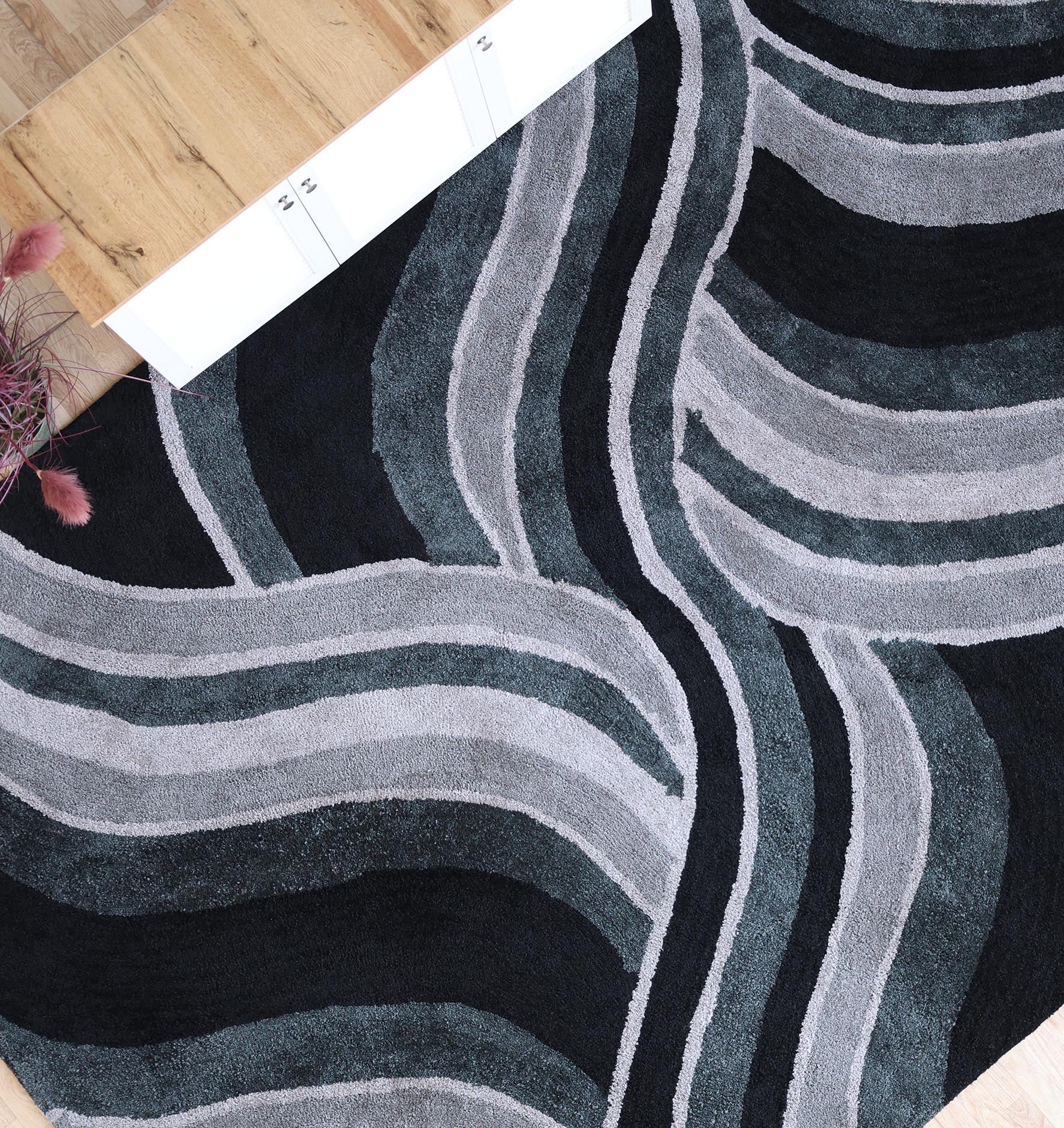 The RAMSHA HOME Multi Shaggy Area Rug for Large Living Room (SG 048) features modern wavy stripes in black, gray, and white, shown on a wooden floor with a color sample strip and a pink decorative plant partially visible nearby.