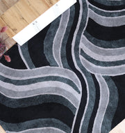 The RAMSHA HOME Multi Shaggy Area Rug for Large Living Room (SG 048) features modern wavy stripes in black, gray, and white, shown on a wooden floor with a color sample strip and a pink decorative plant partially visible nearby.
