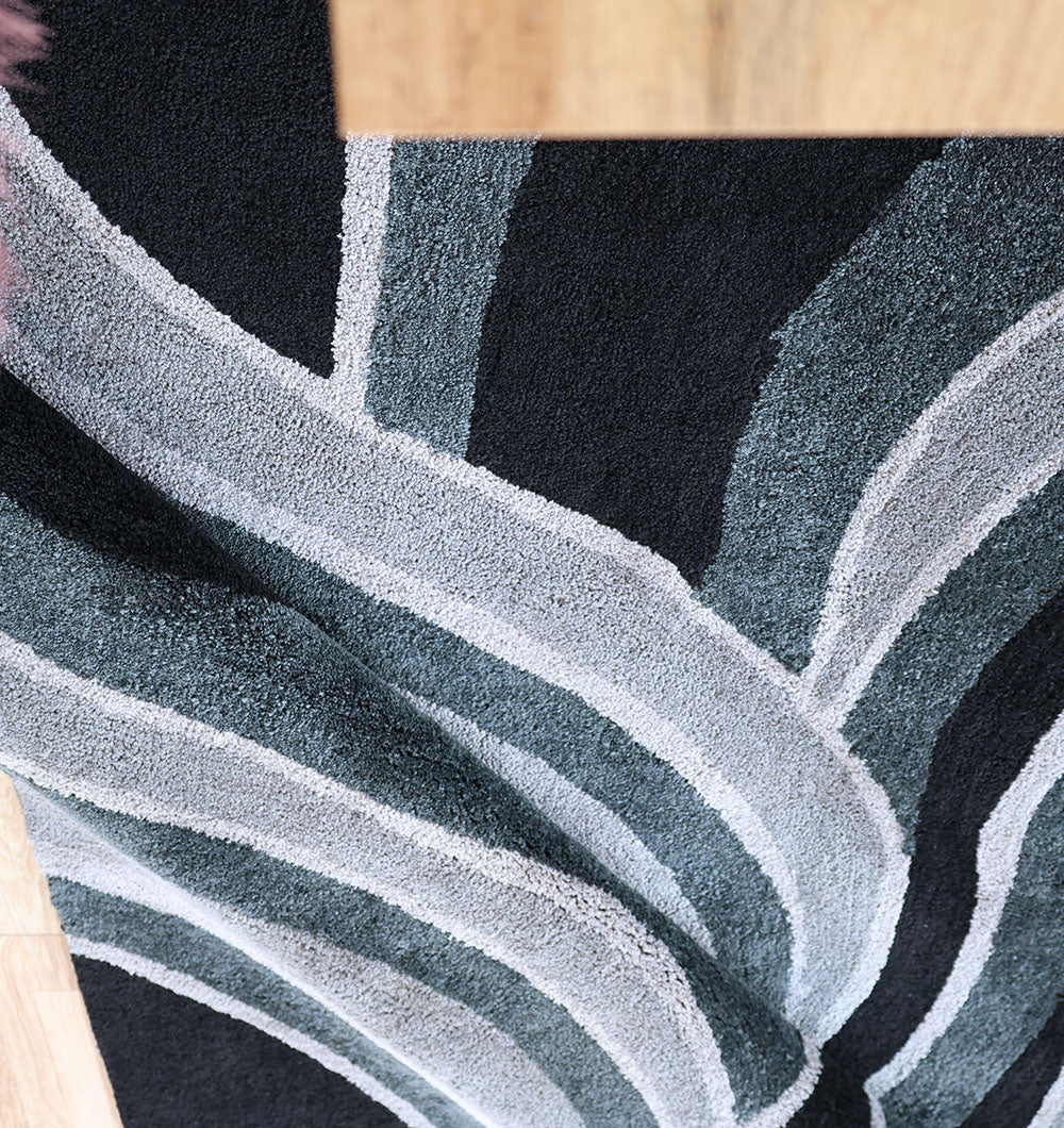 A close-up of the RAMSHA HOME Multi Shaggy Area Rug for Large Living Room (SG 048), featuring plush texture with curved gray and black wavy lines, partially bordered by light wood at the top and bottom.
