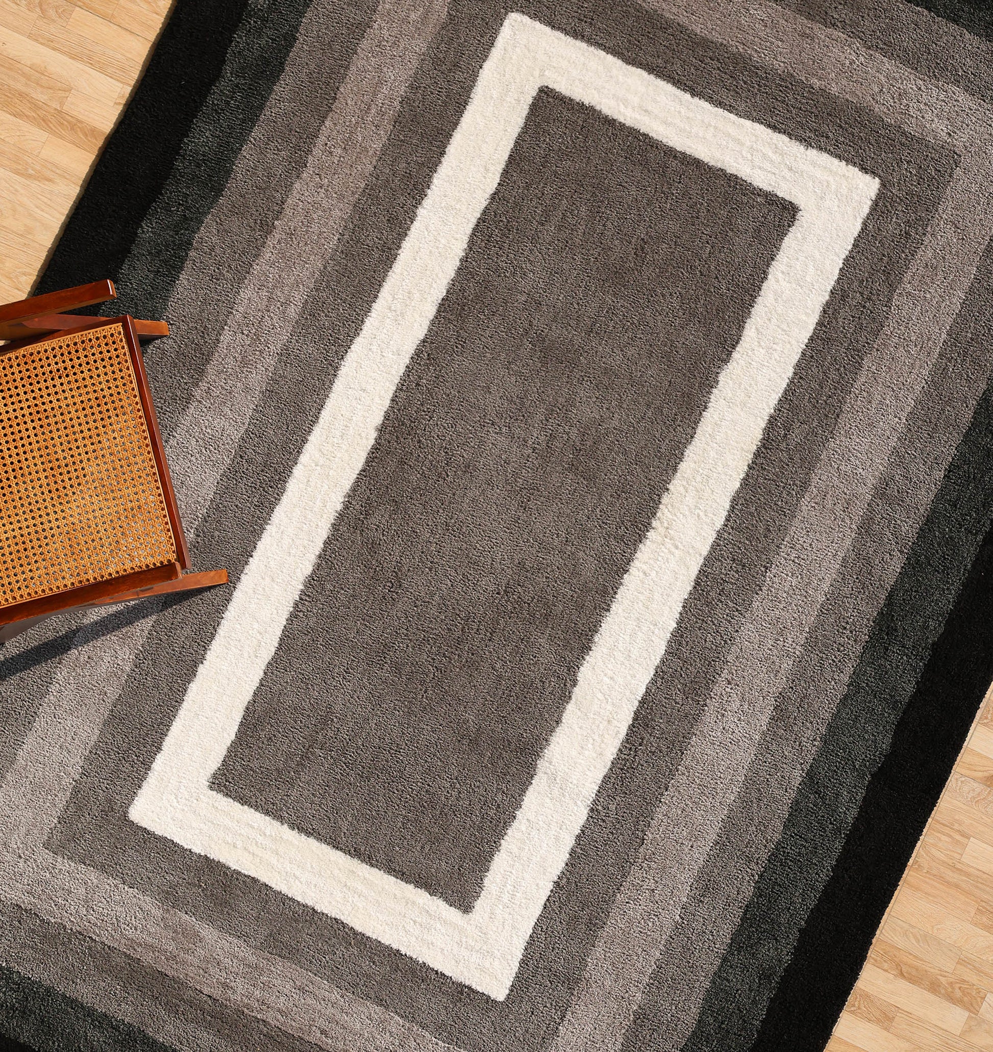 The RAMSHA HOME Black Shaggy Area Rug for Large Living Room (SG 049) with layered gray, white, and black borders sits on a wooden floor. Its plush pile adds comfort, while a wooden chair with a woven cane seat appears in the lower left.