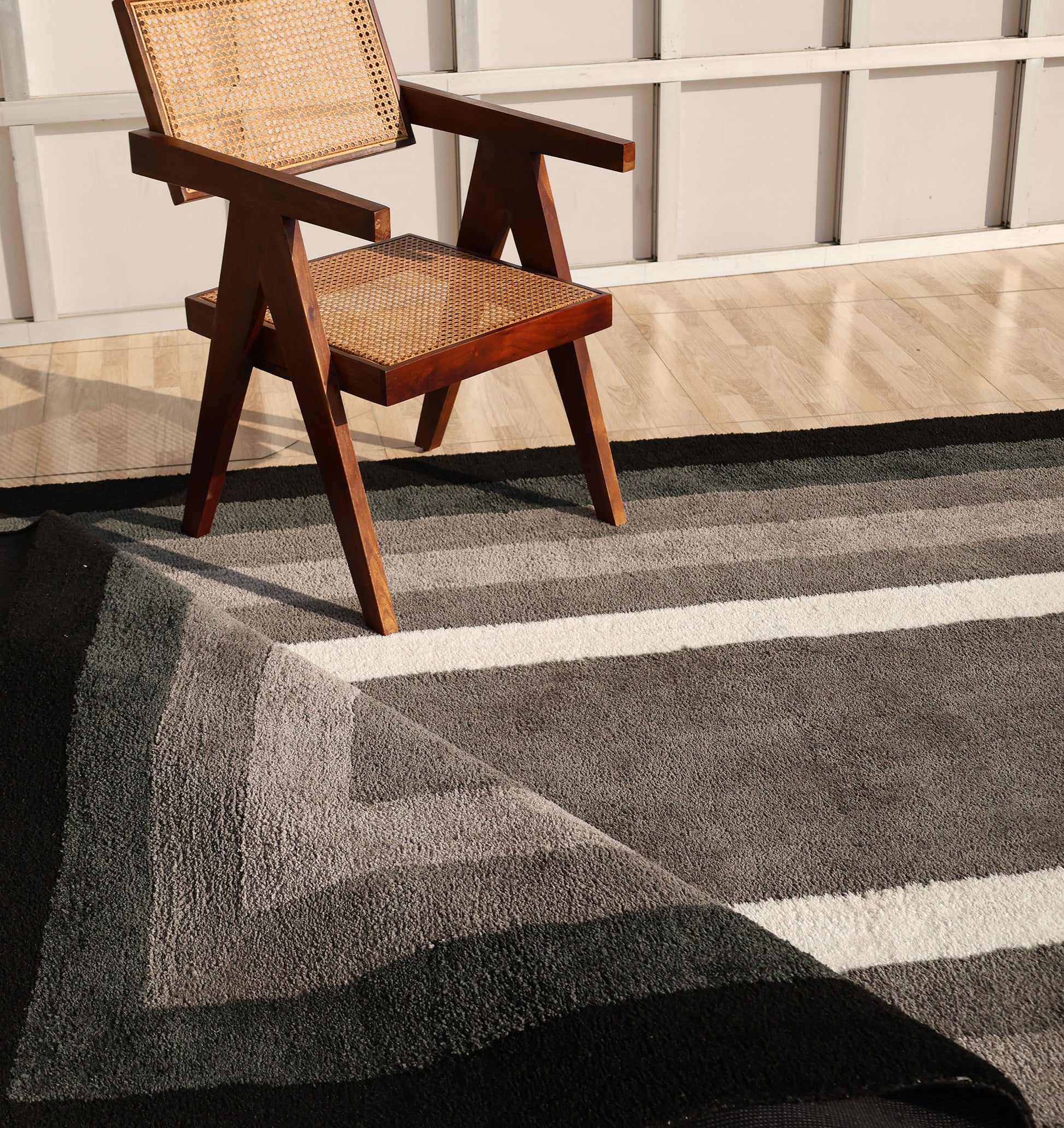 A wooden chair with a woven seat rests on the RAMSHA HOME Black Shaggy Area Rug for Large Living Room (SG 049) in a sunlit space with light wood floors and white paneled walls.