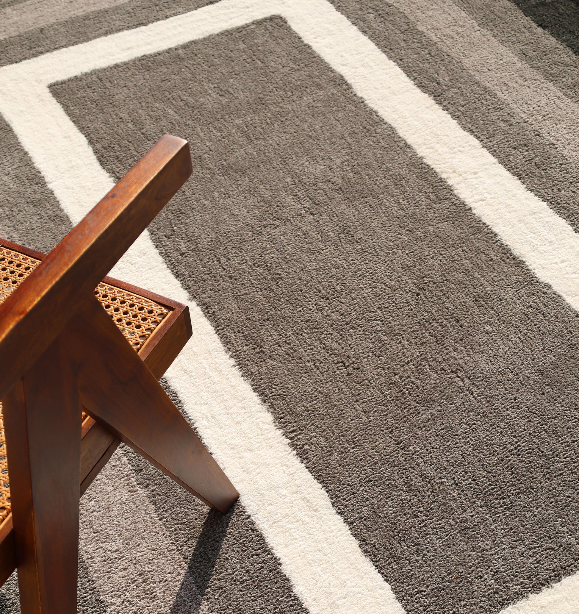 A wooden chair with a woven seat rests on the RAMSHA HOME Black Shaggy Area Rug for Large Living Room (SG 049), its plush pile and geometric gray-and-white pattern bathed in soft sunlight.