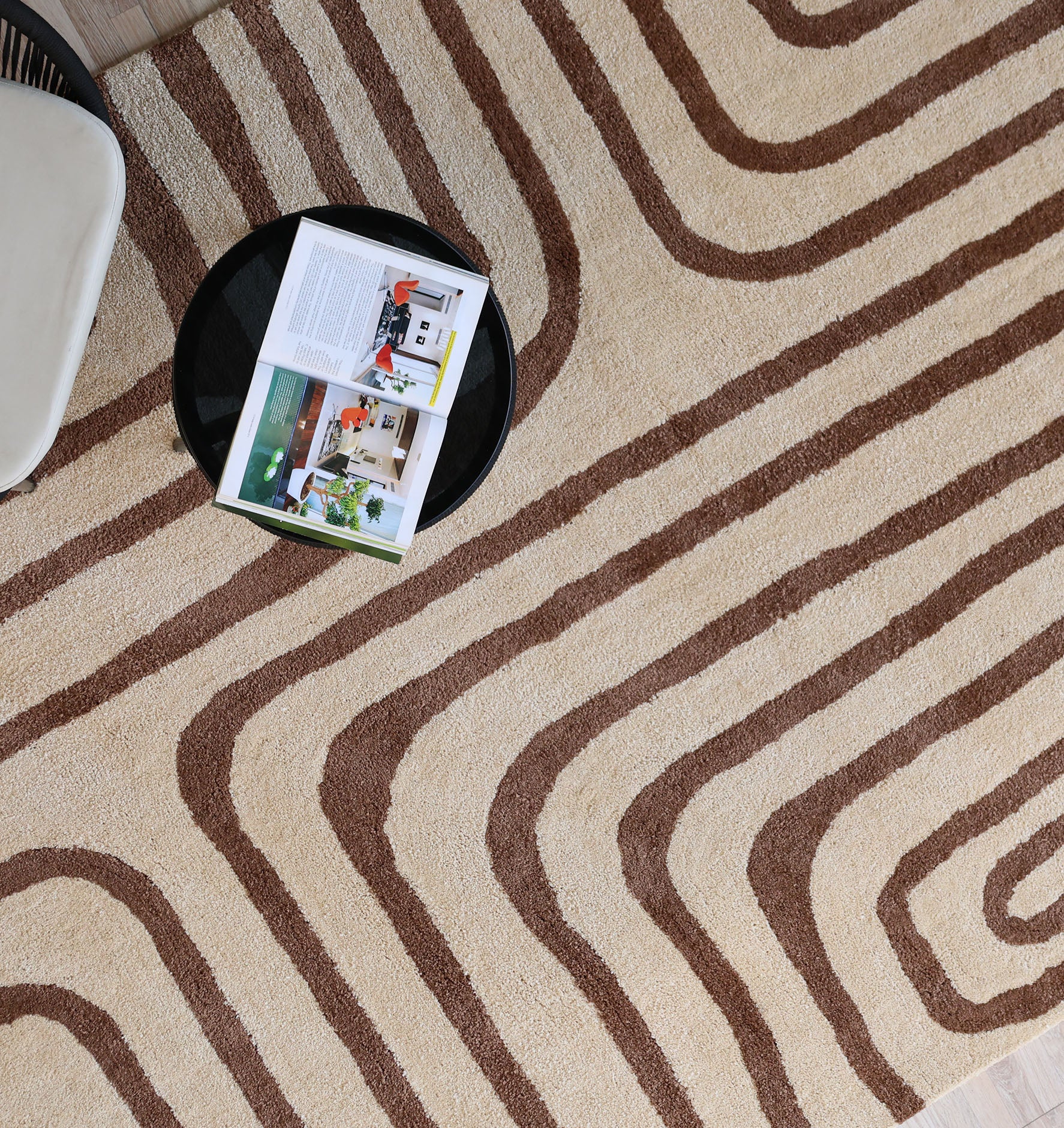 A round black table with an open magazine sits on the RAMSHA HOME Beige and brown Shaggy Area Rug for Large Living Room - SG 050; part of a white chair is visible in the corner.
