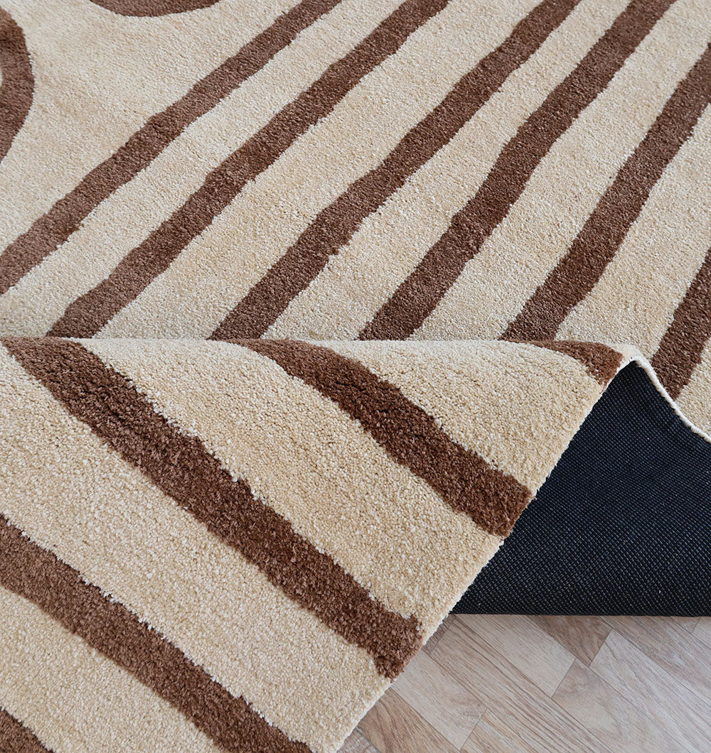 The RAMSHA HOME Beige and Brown Shaggy Area Rug for Large Living Room (SG 050) features plush beige fibers with brown stripes, shown partially lifted at the corner to reveal a black non-slip rug pad on a light wood floor.