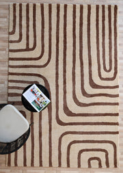 The RAMSHA HOME Beige and Brown Shaggy Area Rug for Large Living Room - SG 050 lies on a wooden floor beside a modern chair with a light cushion and a round black side table holding an open magazine.