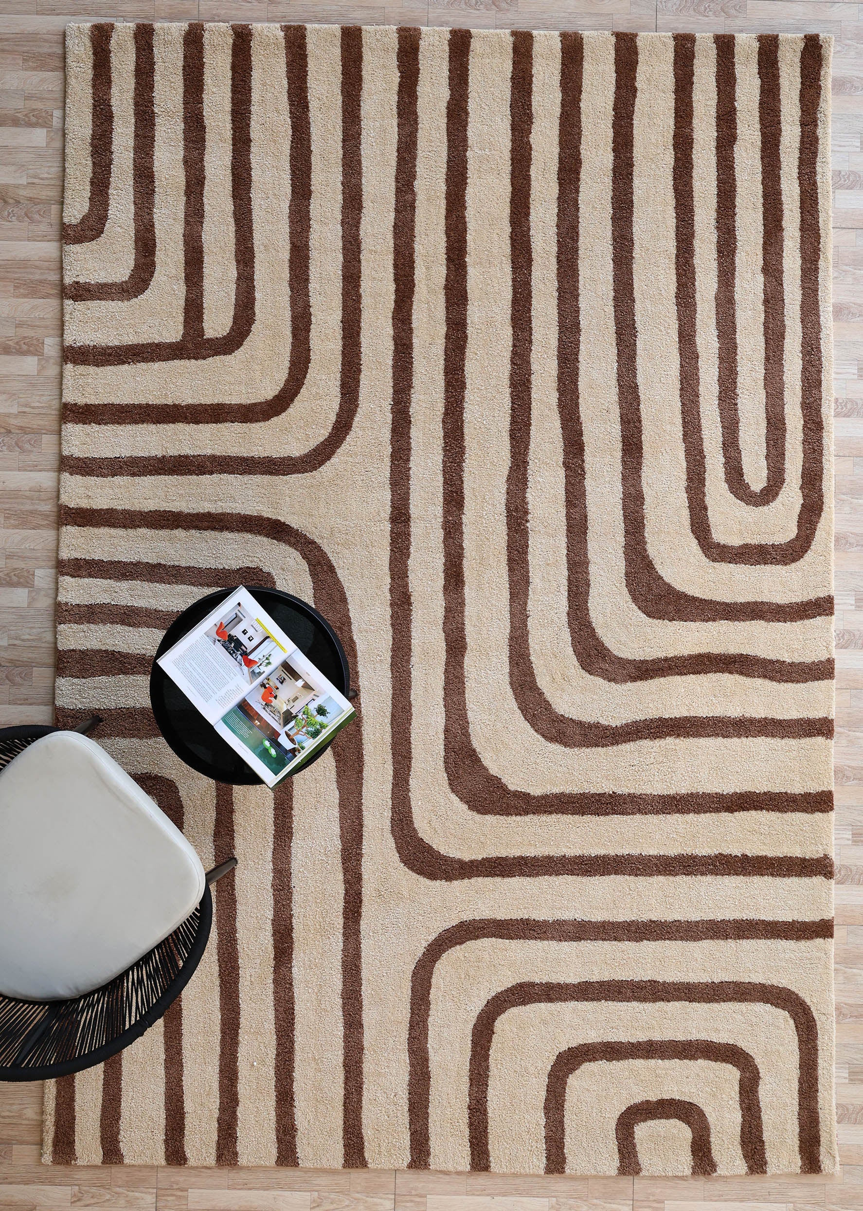 The RAMSHA HOME Beige and Brown Shaggy Area Rug for Large Living Room - SG 050 lies on a wooden floor beside a modern chair with a light cushion and a round black side table holding an open magazine.