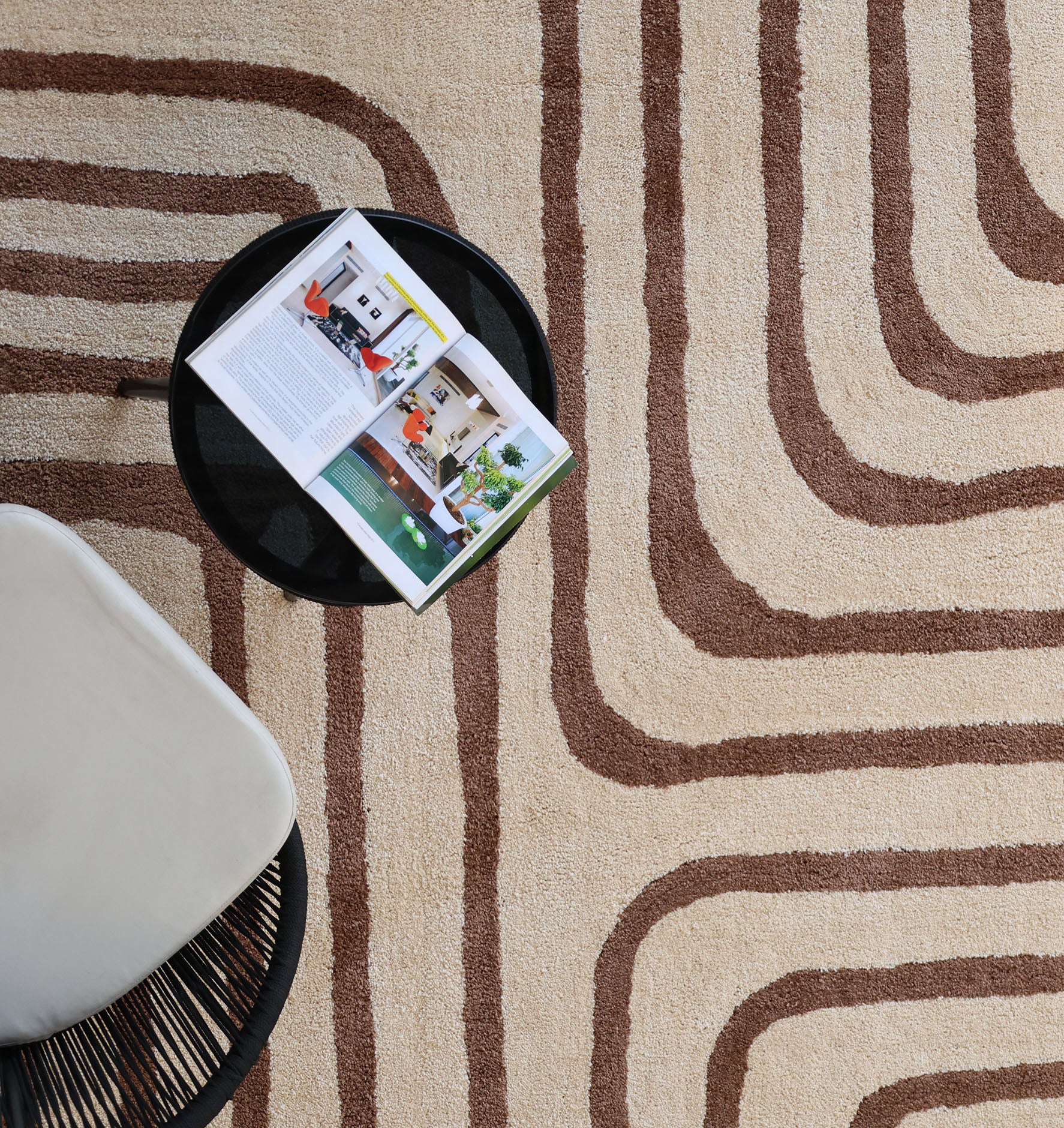 A round black side table with an open magazine is next to a white chair on the RAMSHA HOME Beige and brown Shaggy Area Rug for Large Living Room - SG 050, featuring plush pile and curved brown geometric lines.