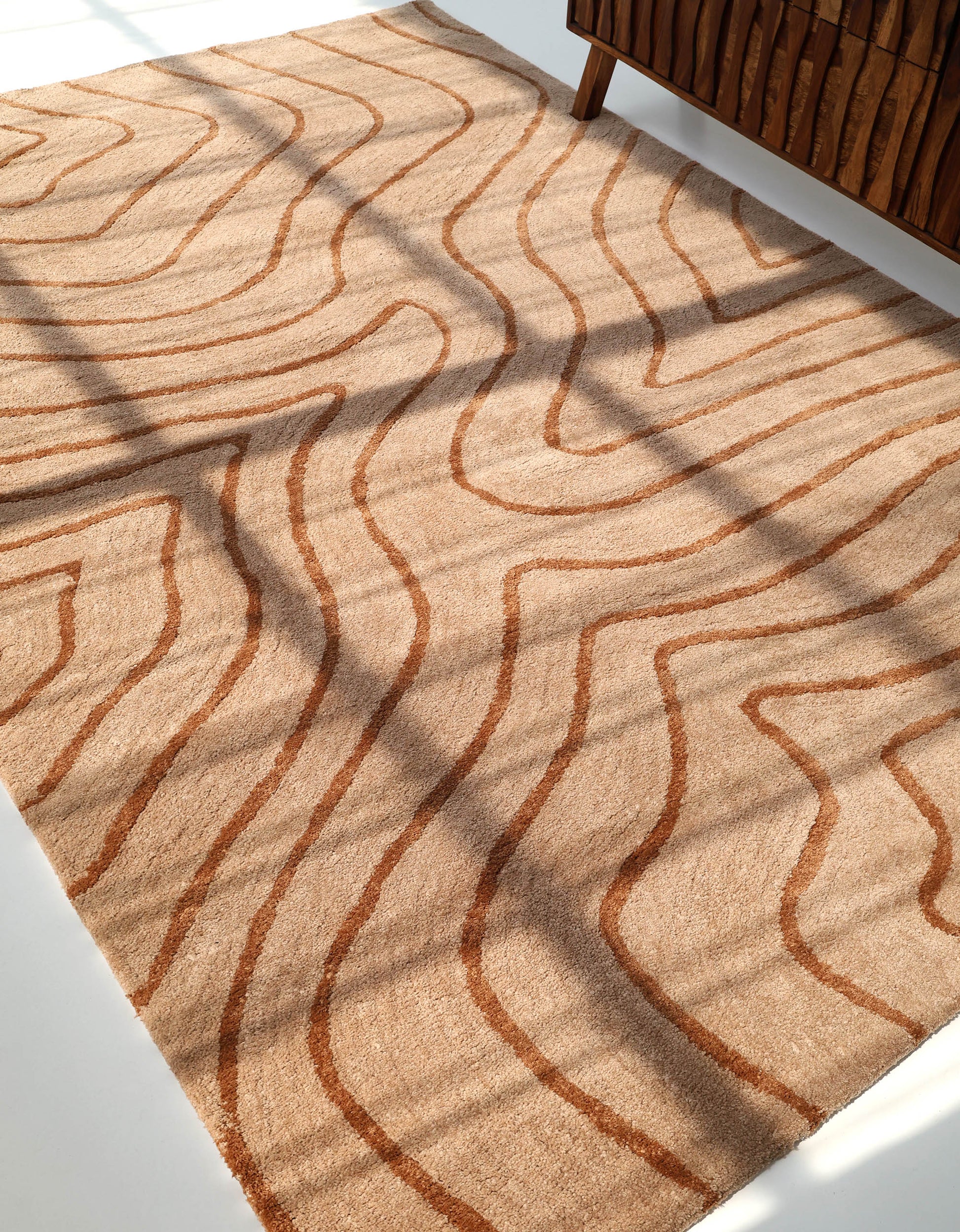 The RAMSHA HOME Designer Contour Line Plush Area Rug - SG 051 features modern beige-brown tones and rust wavy lines forming an abstract pattern, shown sunlit with shadows and a wooden cabinet in the top right corner.