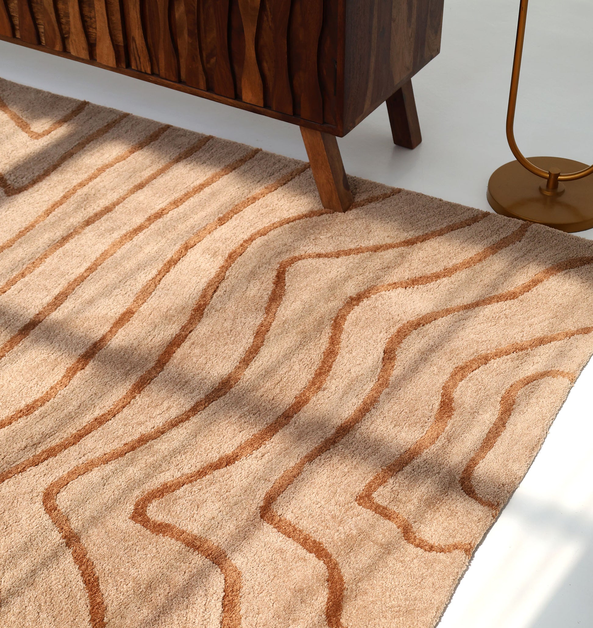 The RAMSHA HOME Designer Contour Line Plush Area Rug - SG 051, featuring wavy beige and brown lines, sits on a white floor beside a textured wood cabinet and gold lamp base, with sunlight casting elegant shadows across its plush surface.