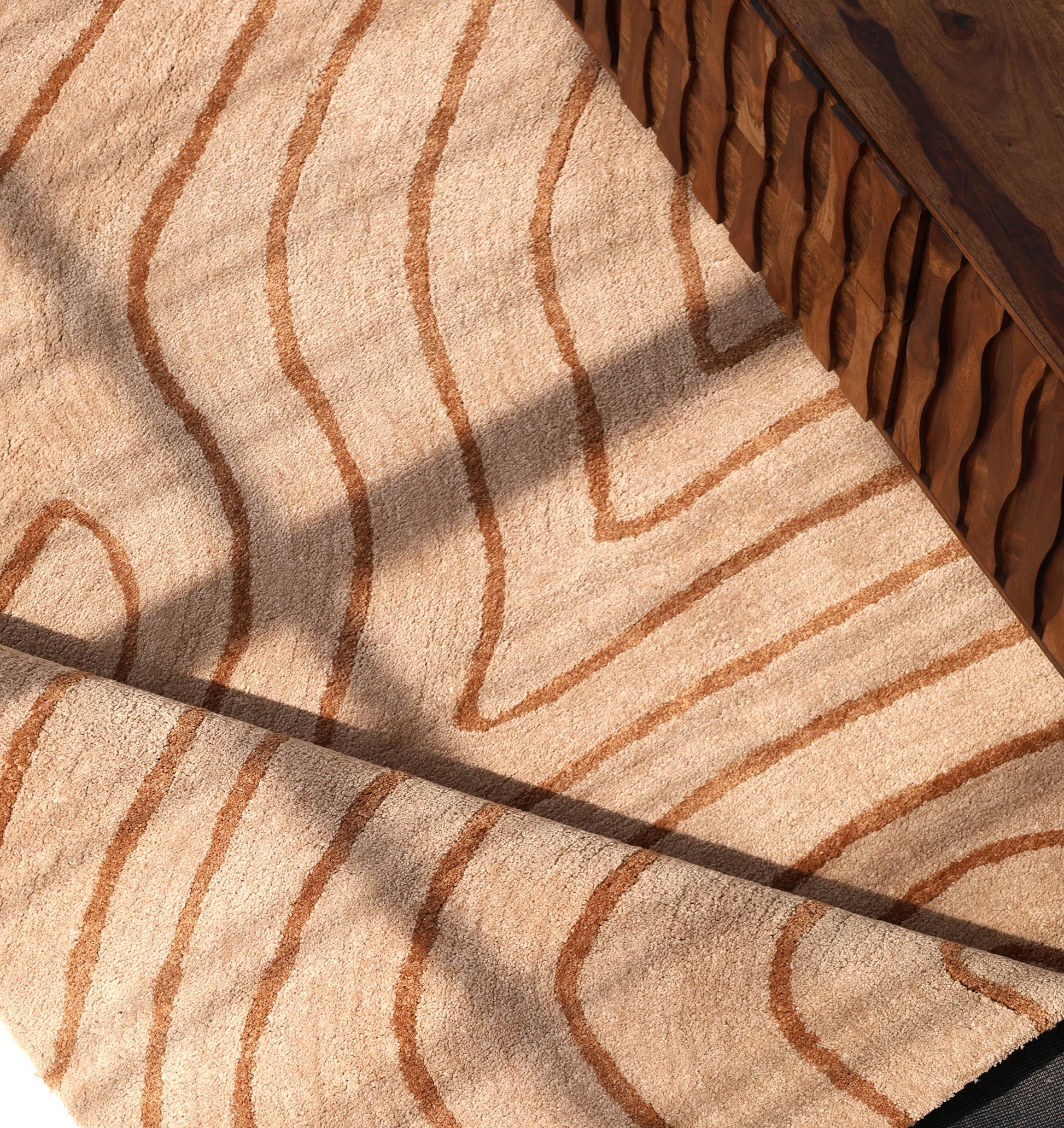 The RAMSHA HOME Designer Contour Line Plush Area Rug - SG 051, featuring wavy wood-grain patterns in beige and brown, lies beside a carved-edge wooden table as sunlight highlights its unique texture and abstract design.
