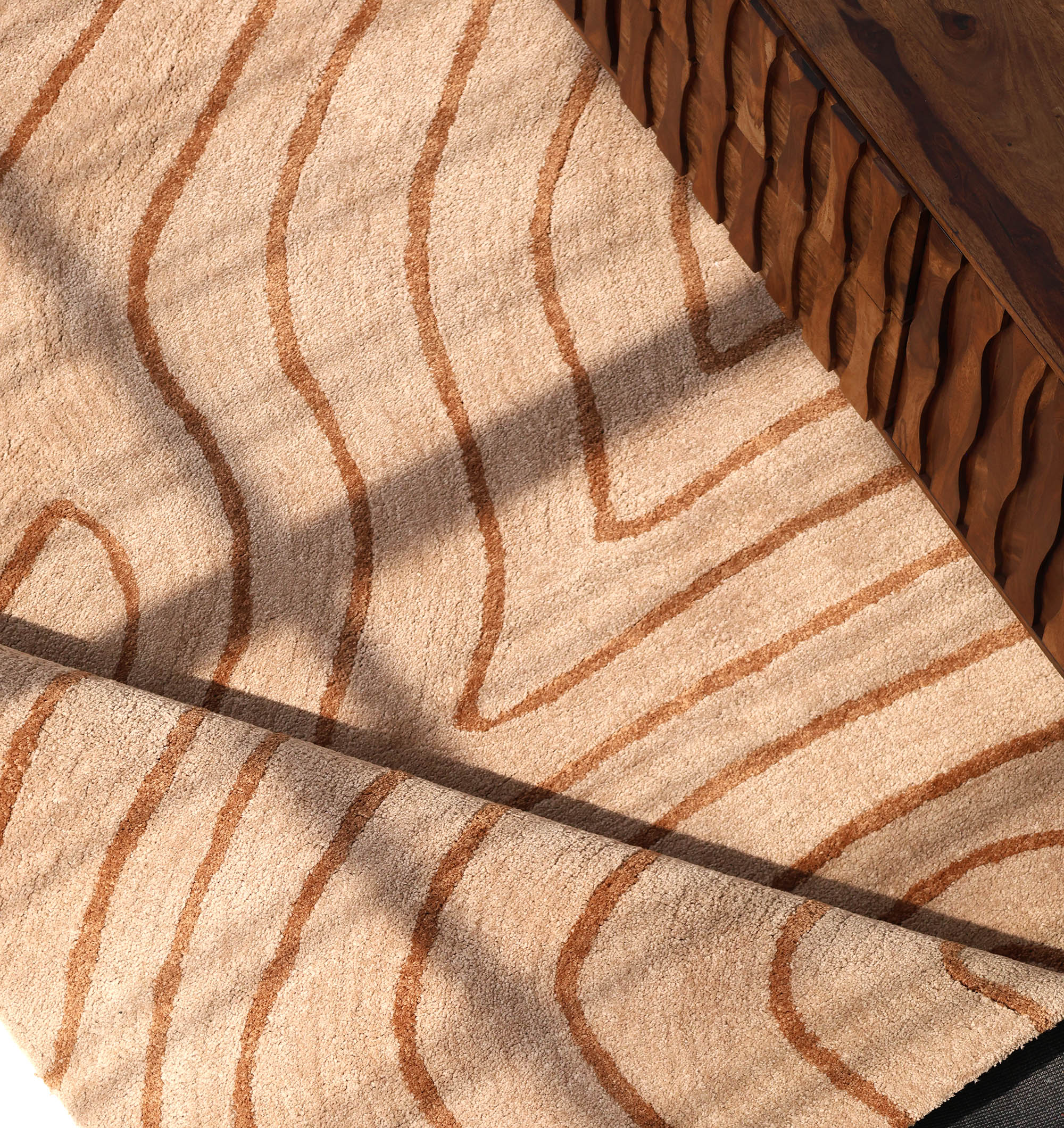 The RAMSHA HOME Designer Contour Line Plush Area Rug - SG 051, featuring wavy wood-grain patterns in beige and brown, lies beside a carved-edge wooden table as sunlight highlights its unique texture and abstract design.