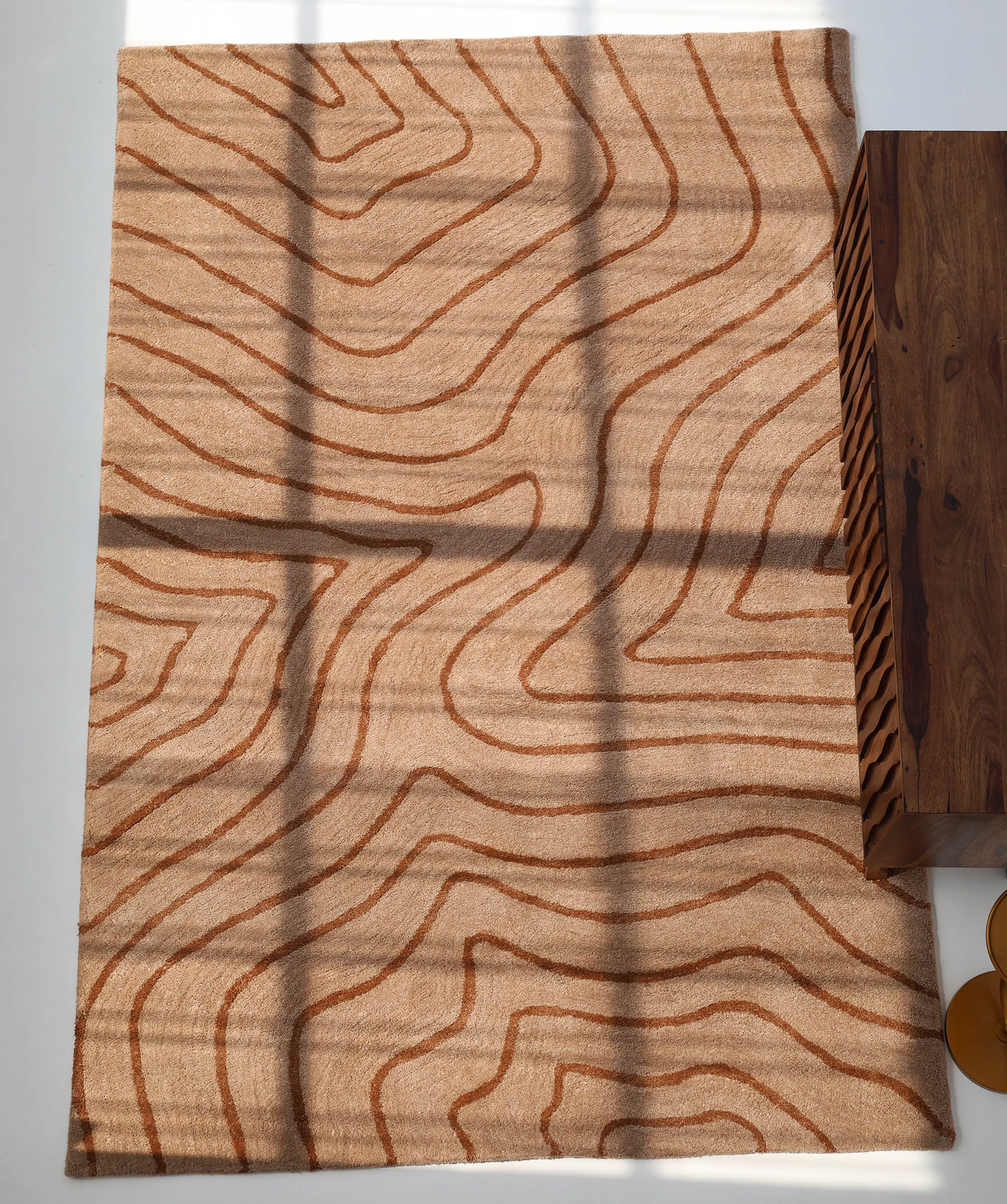The RAMSHA HOME Designer Contour Line Plush Area Rug - SG 051, in beige brown with abstract rust wavy patterns, is partially sunlit next to a wooden table on a white floor.