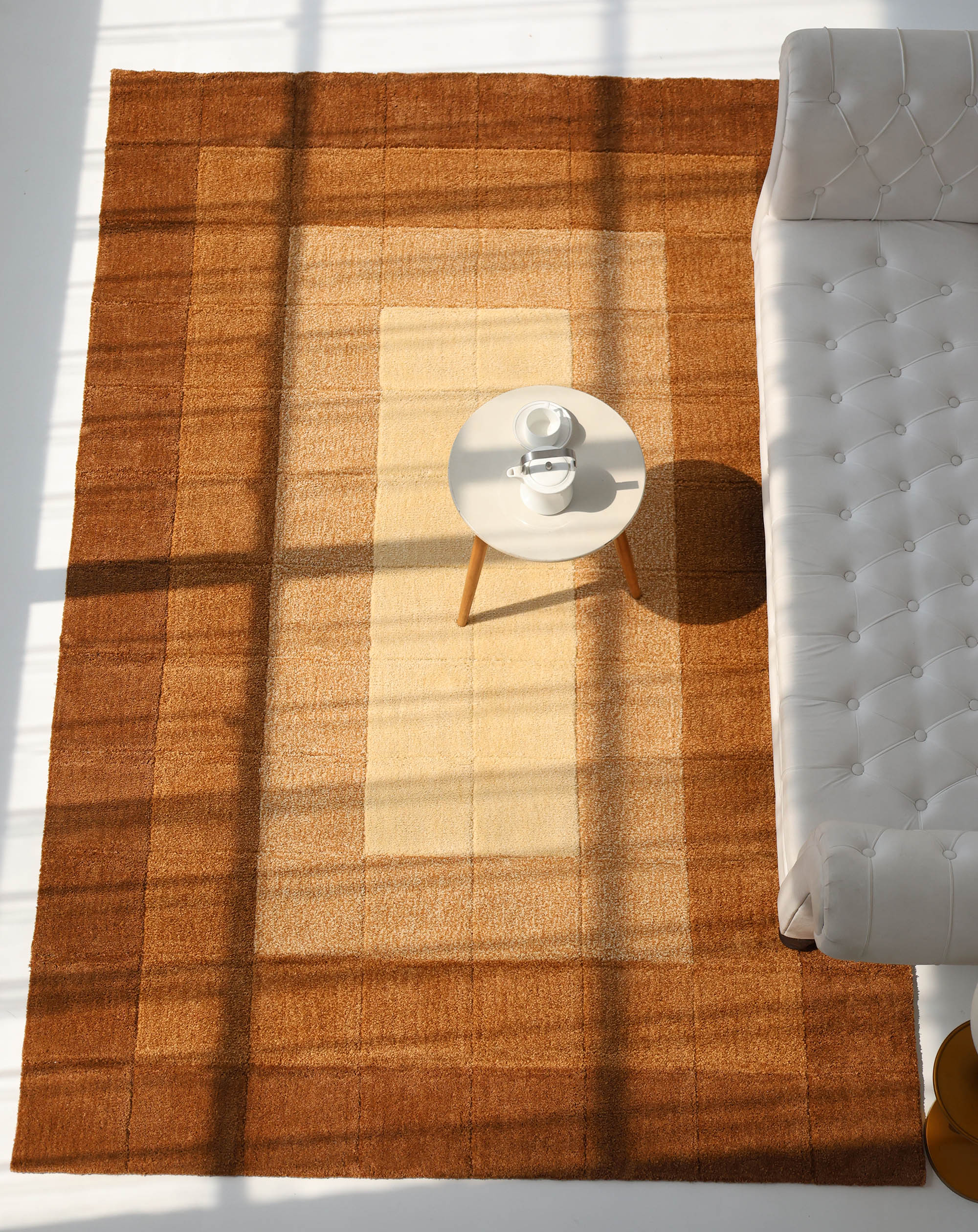 The RAMSHA HOME Modern Beige & Brown circular Shaggy Rug for Living Room - SG 052 lies under a small round white table with cups and saucers beside a white tufted sofa, as sunlight and window shadows sweep across its subtle gradient pattern.