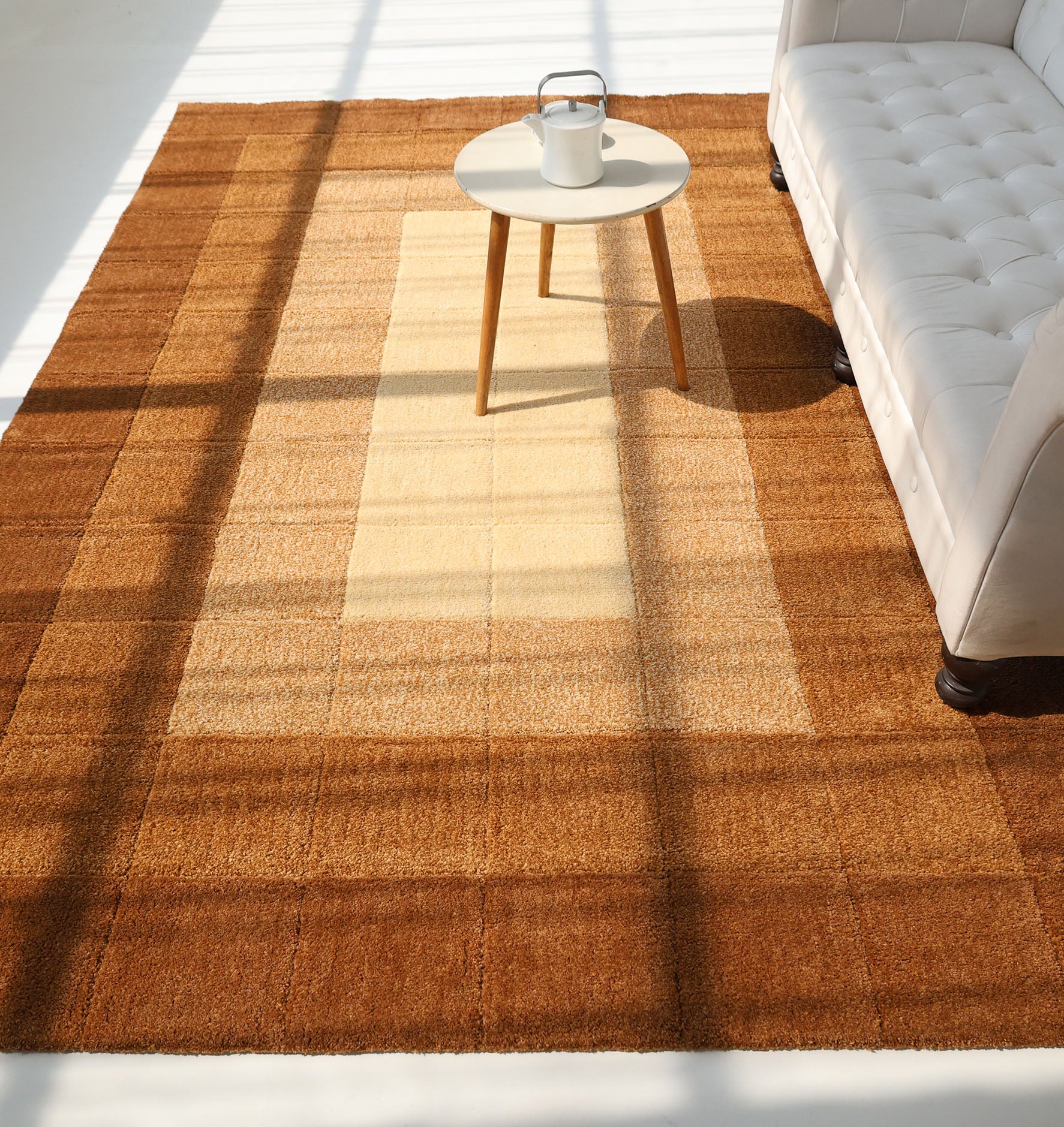 The RAMSHA HOME Modern Beige & Brown circular Shaggy Rug (SG 052) adds warmth to the sunlit room, paired with a white round side table and a light beige tufted sofa as sunlight casts windowpane shadows across the cozy setting.