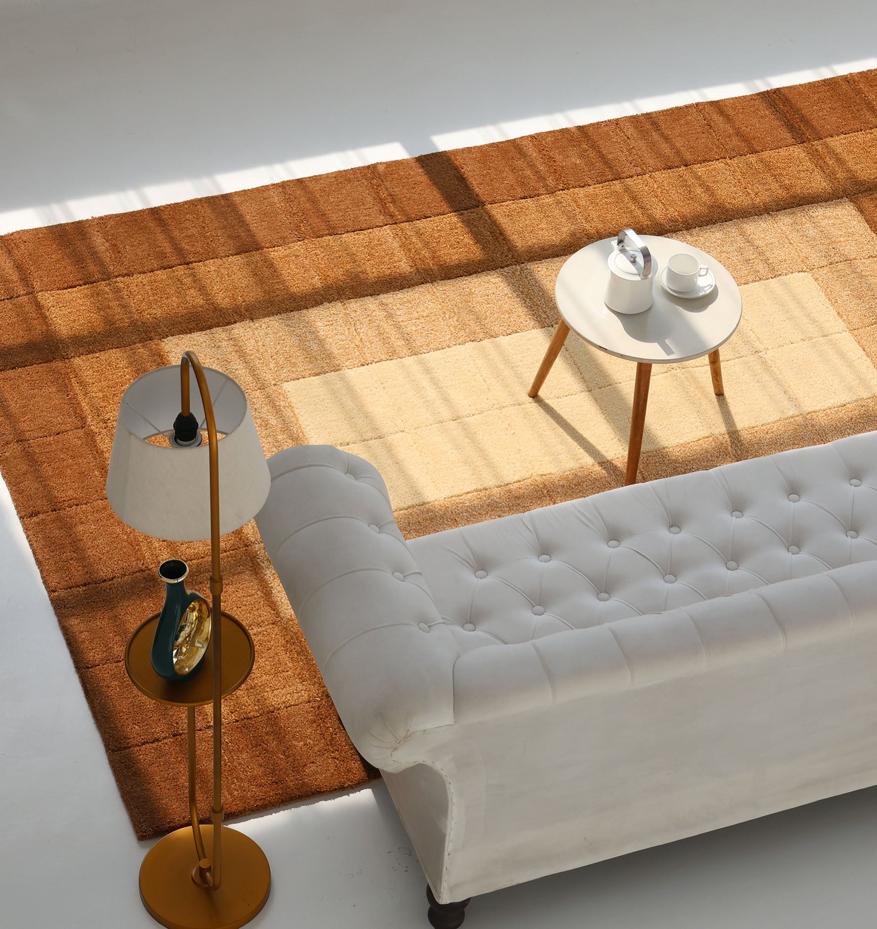 A cozy living room with a white tufted sofa, round white side table set with teapot and cups, and a RAMSHA HOME Modern Beige & Brown circular Shaggy Rug for Living Room (Model: SG 052), all bathed in sunlight from large windows.